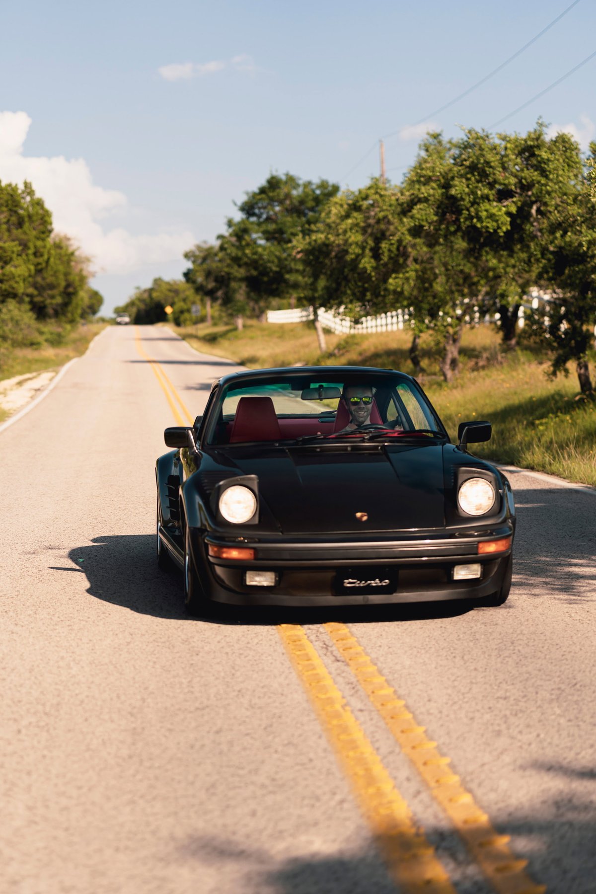 Black 1989 Porsche 930 Turbo Flachbau with red interior driving on a rural road.