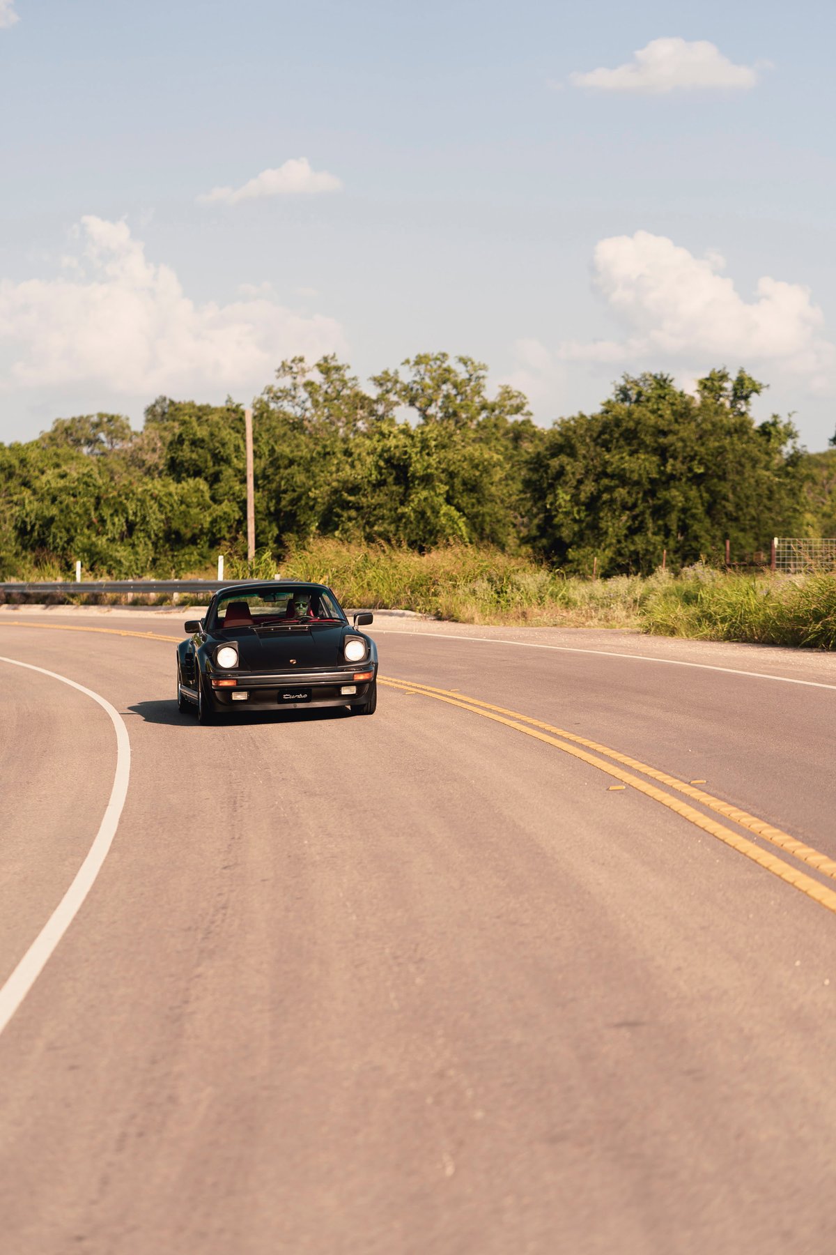 Black 1989 Porsche 930 Turbo Flachbau driving on a rural road.