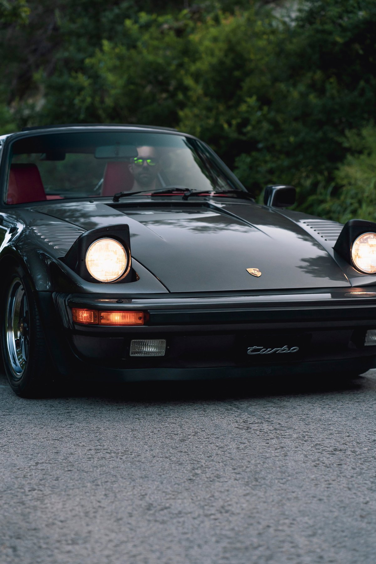 1989 Porsche 930 Turbo Flachbau in dark gray. Pop-up headlights, red interior, "Turbo" script.