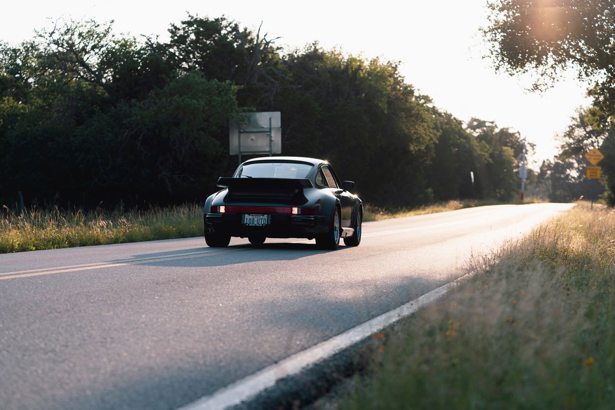 Black 1989 Porsche 930 Turbo Flachbau driving on a road with a rear spoiler.