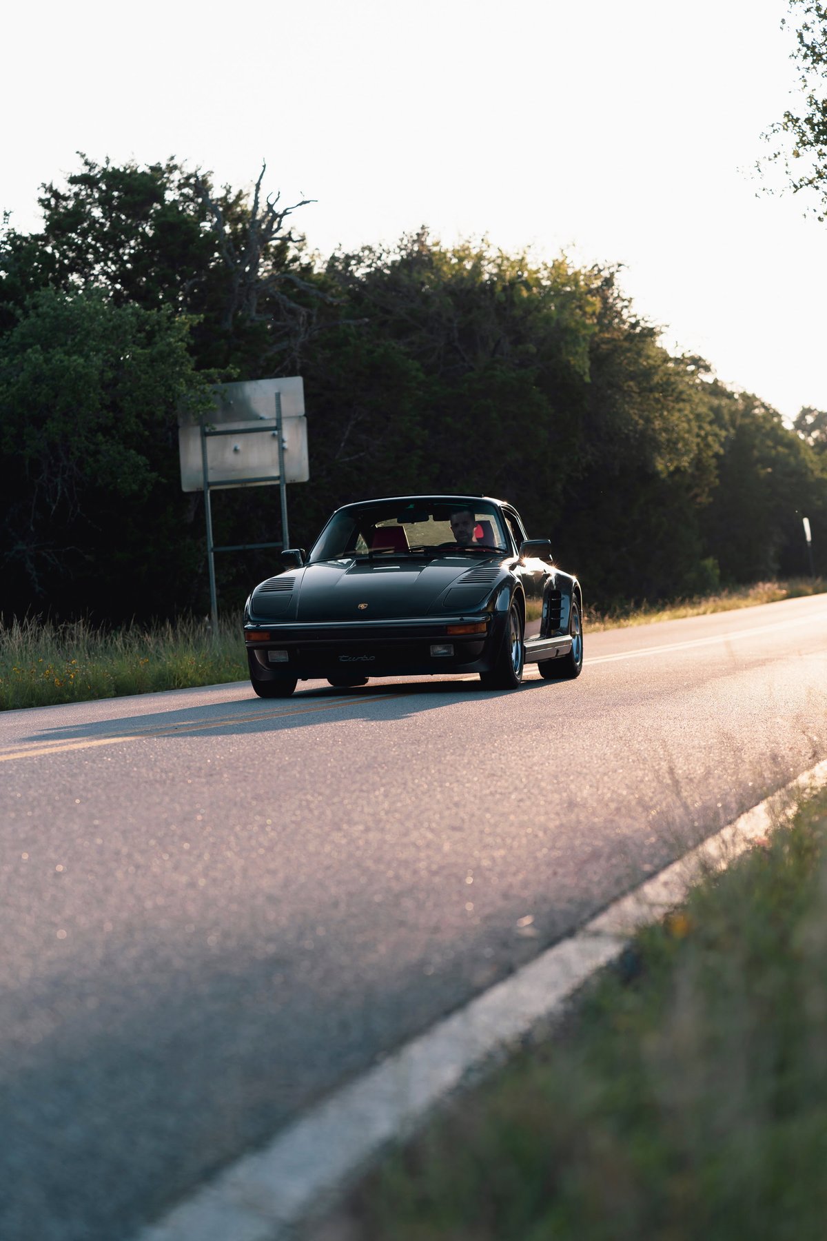 Black 1989 Porsche 930 Turbo Flachbau coupe driving on a road at sunset.