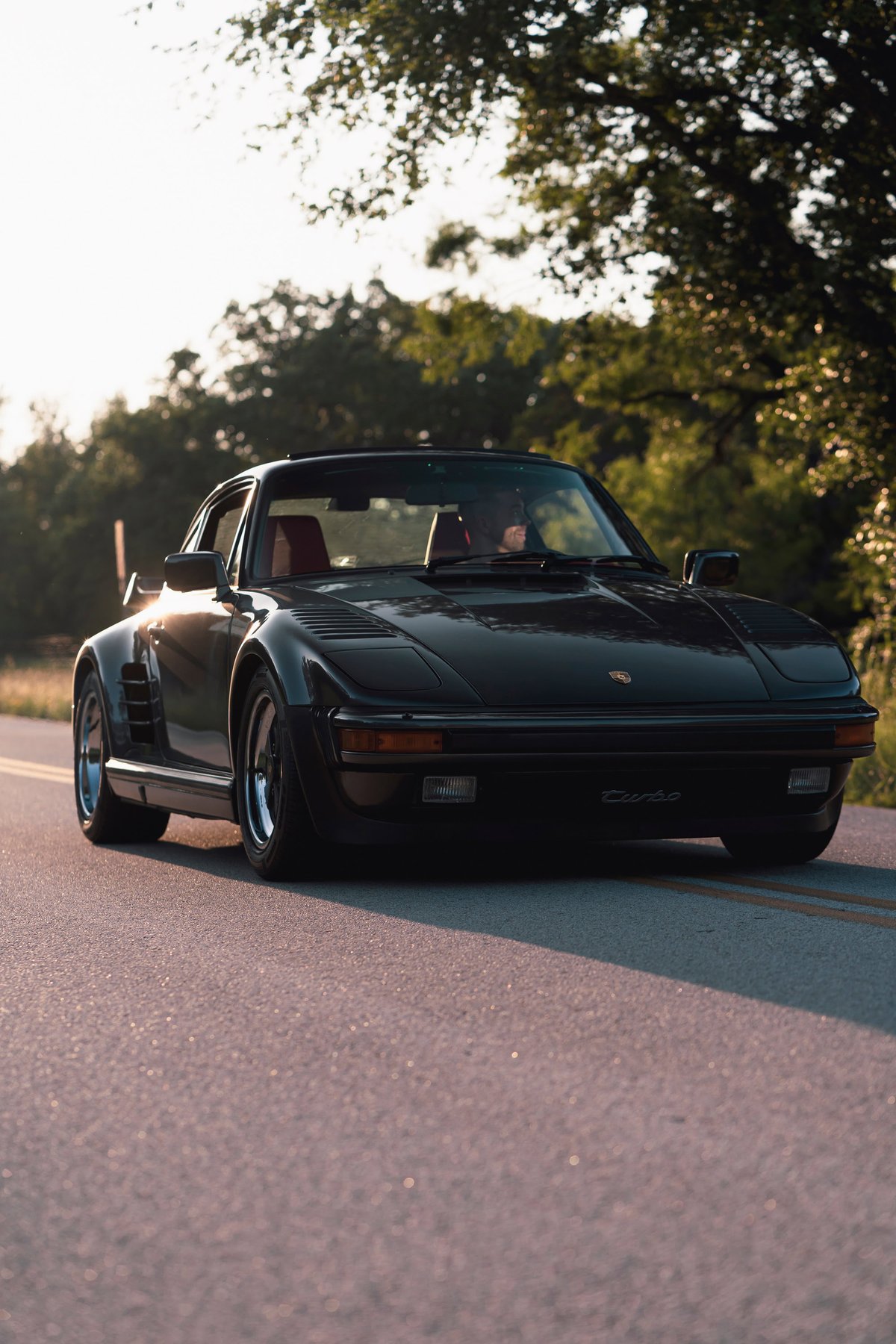 Black 1989 Porsche 930 Turbo Flachbau on road, showing its wide body kit and signature rims.