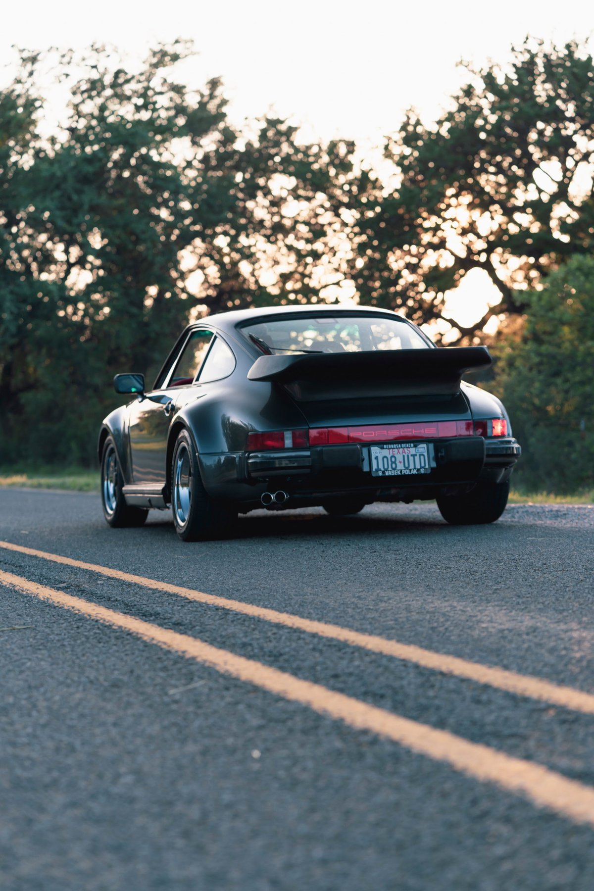 Black 1989 Porsche 930 Turbo Flachbau with whale tail spoiler, polished rims, and dual exhaust.