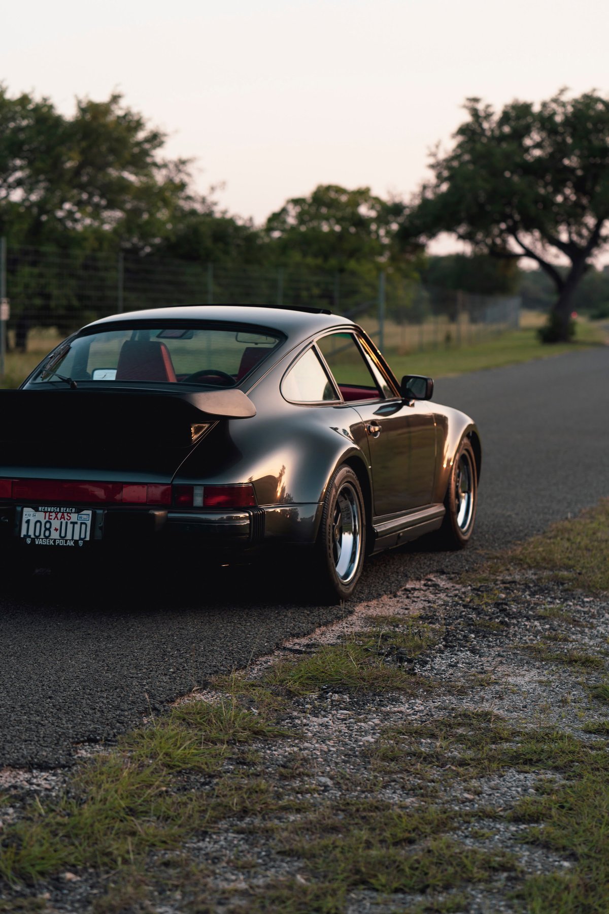1989 Porsche 930 Turbo Flachbau in black with red interior, showing its signature whale tail spoiler and classic rims.