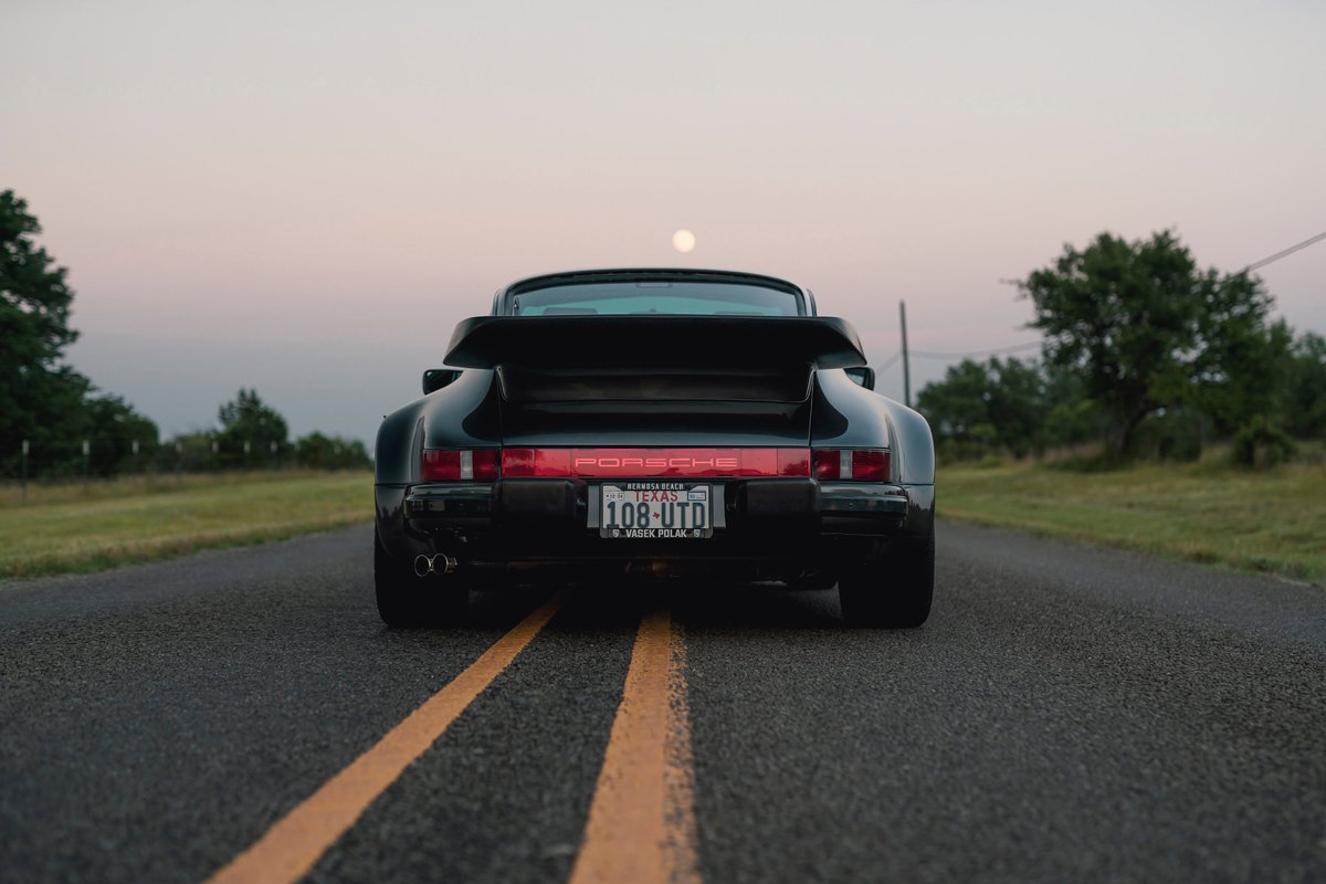 Rear view of a black 1989 Porsche 930 Turbo Flachbau on a road at dusk, showing its wide body kit and whale tail spoiler.