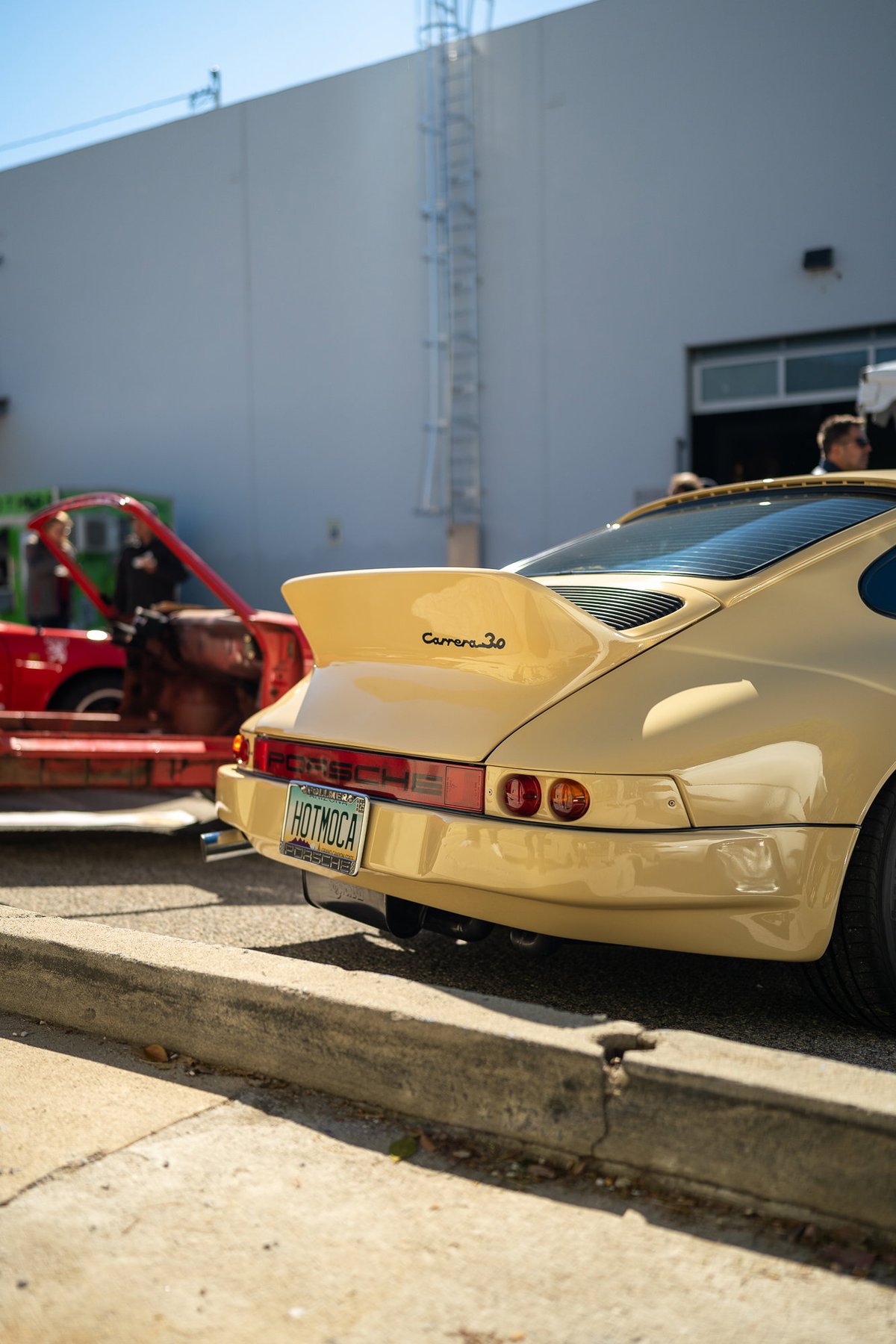 Rear view of a tan Porsche 911 Carrera 3.0 with a ducktail spoiler and "HOTMOCA" license plate.