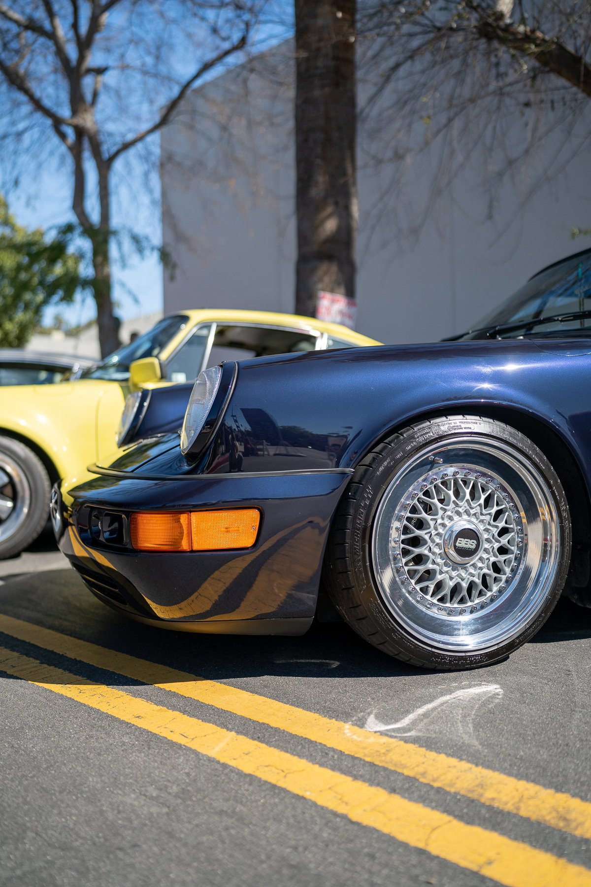 Close-up of a dark blue Porsche 911 with silver BBS rims and a yellow Porsche 911 in the background.
