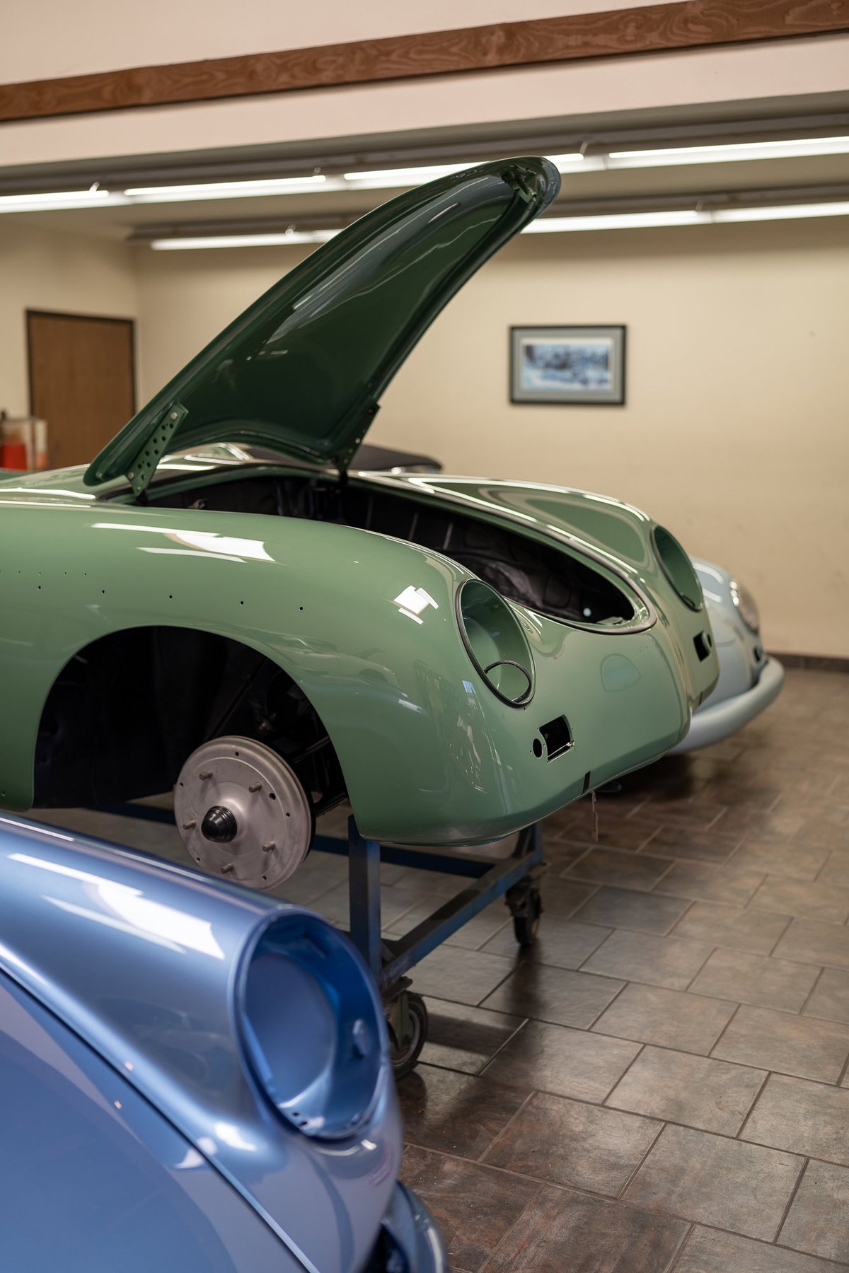 Vintage light green Porsche 356, hood open, bare rims, in shop.