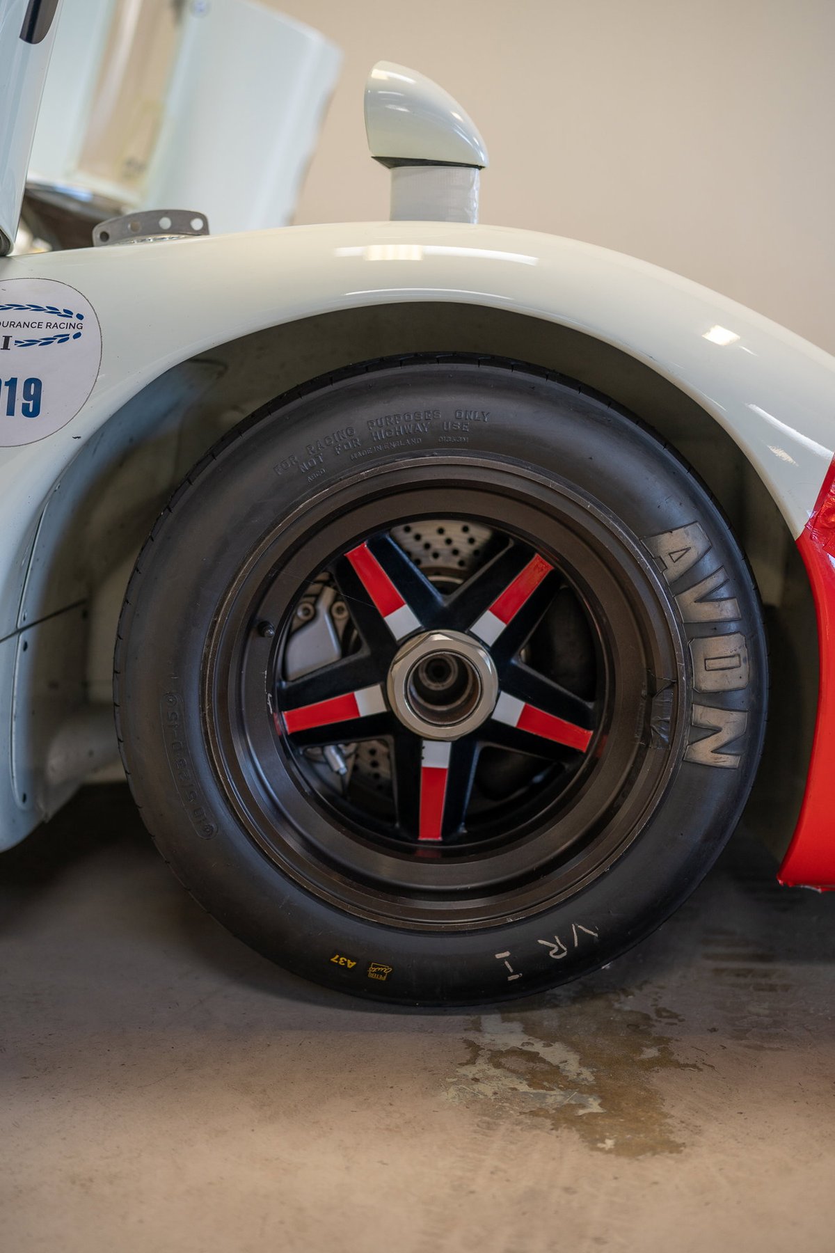 Close-up of a white Porsche 911 race car's wheel, featuring a black 5-star rim with red and silver accents, and Avon tires.