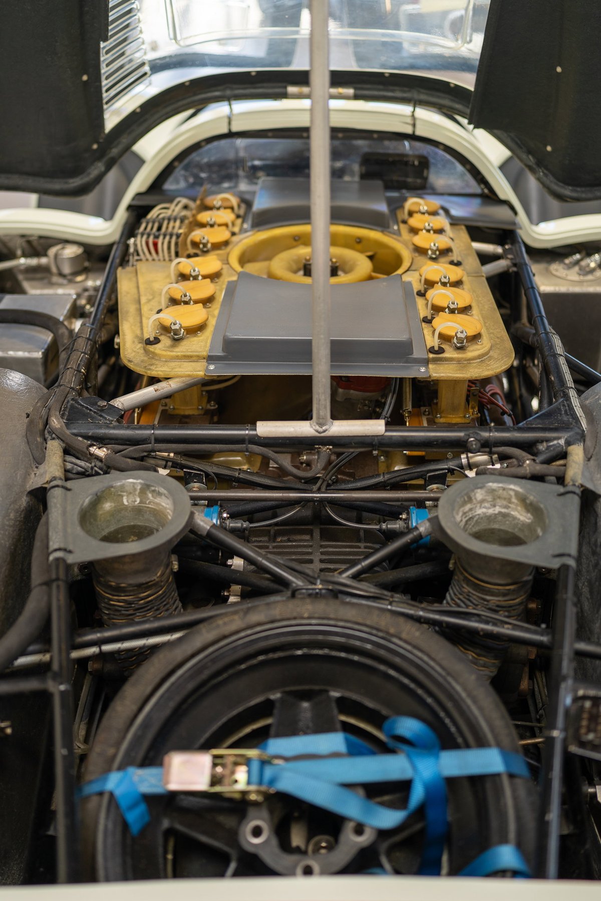 Vintage race car engine bay, exposed V10, exposed yellow intake trumpets, and a spare tire secured with blue straps.