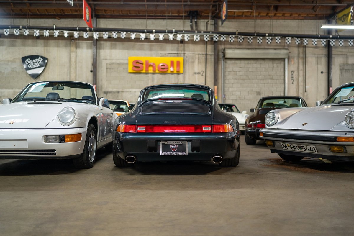 Rear view of a dark green Porsche 911 Carrera in a showroom, with other Porsches visible.