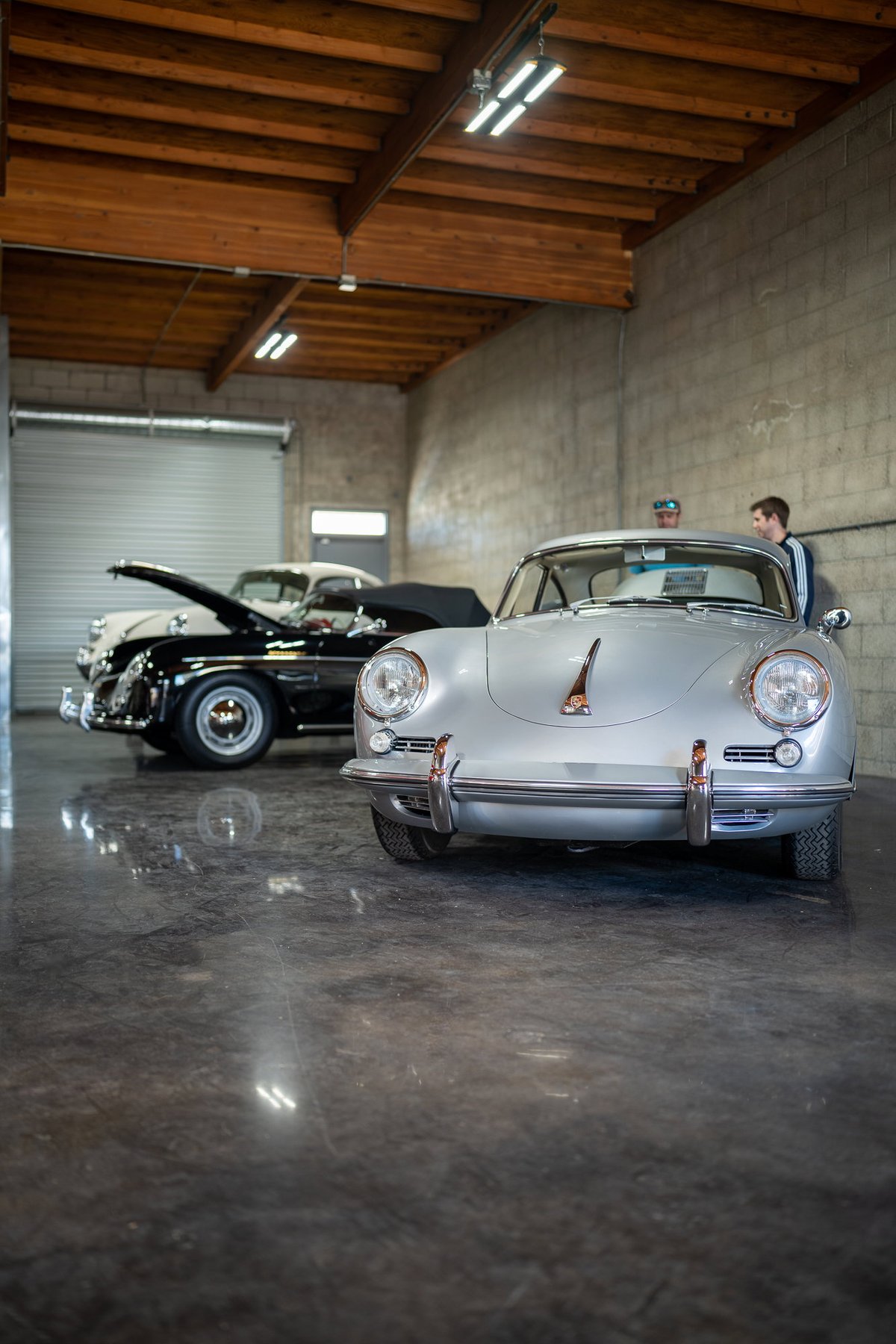 Silver Porsche 356 Carrera 2 coupe in a garage with a black convertible in the background.