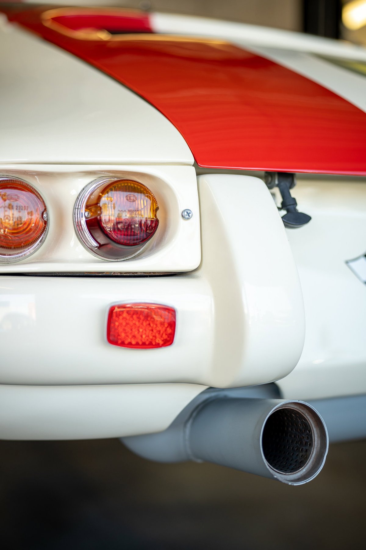 Close-up of a vintage white sports car's rear, showing dual red taillights, a red stripe livery, and a chrome exhaust pipe.