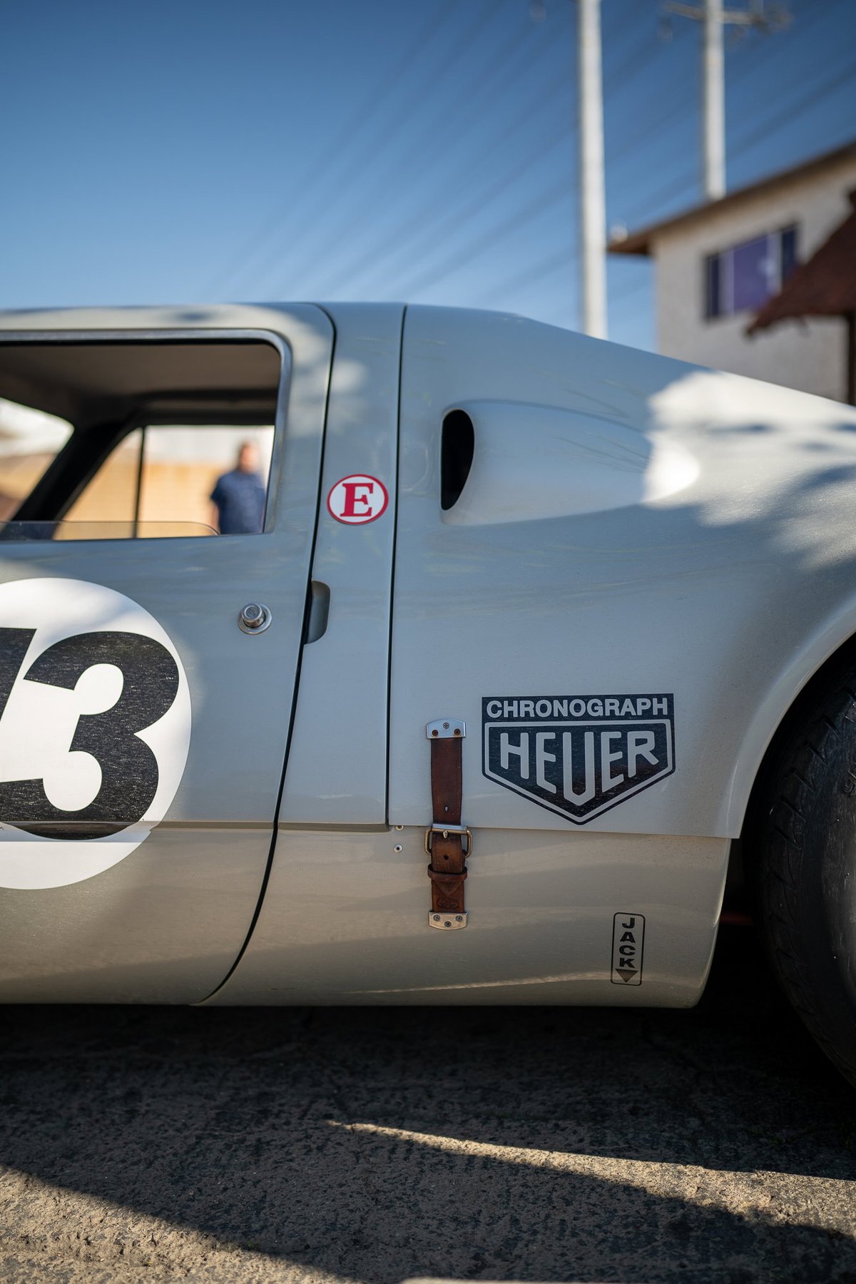Close-up of a classic race car, possibly a Porsche, in light grey. Features a large "73" racing number and a Heuer Chronograph badge.