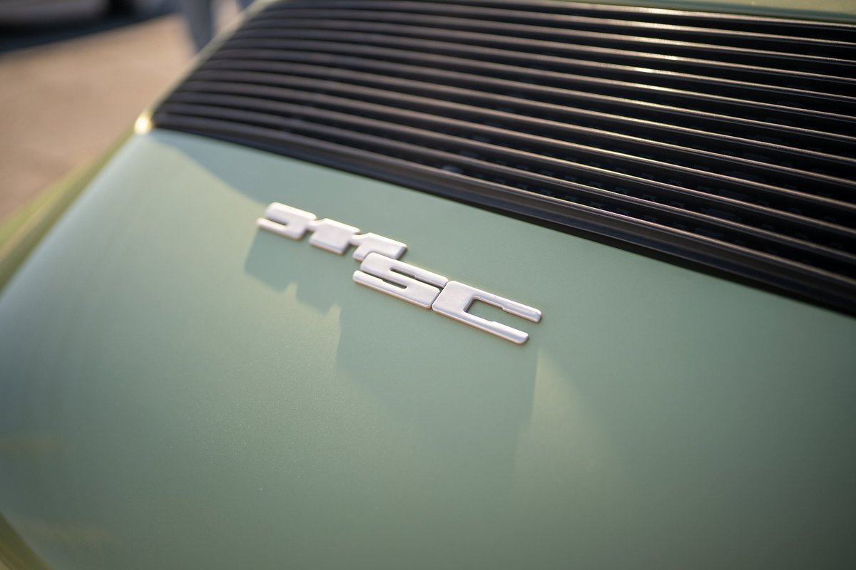 Close up of a Porsche 911 SC badge in silver on a pale green rear deck lid with black louvres.