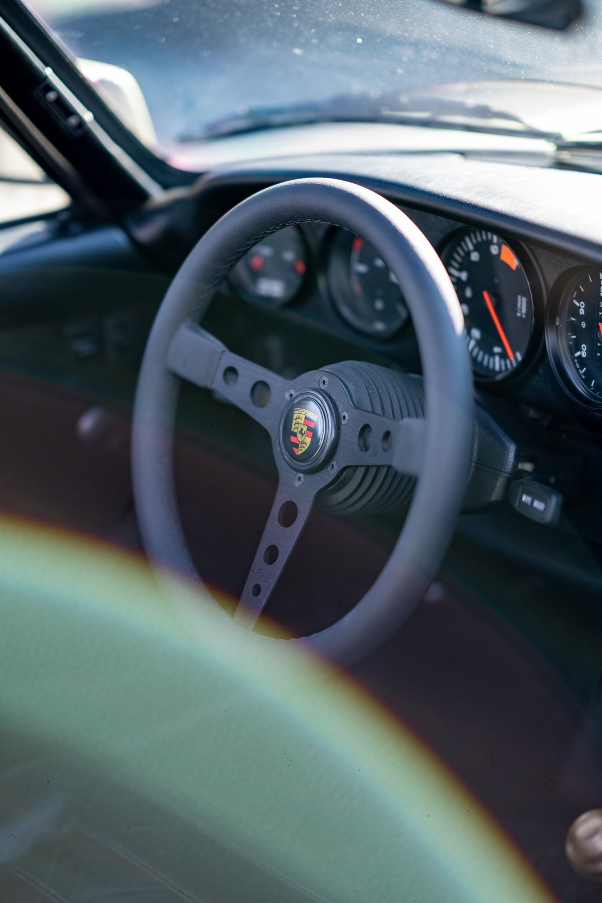 Interior view of a black Porsche steering wheel with the Porsche crest and a vintage tachometer.