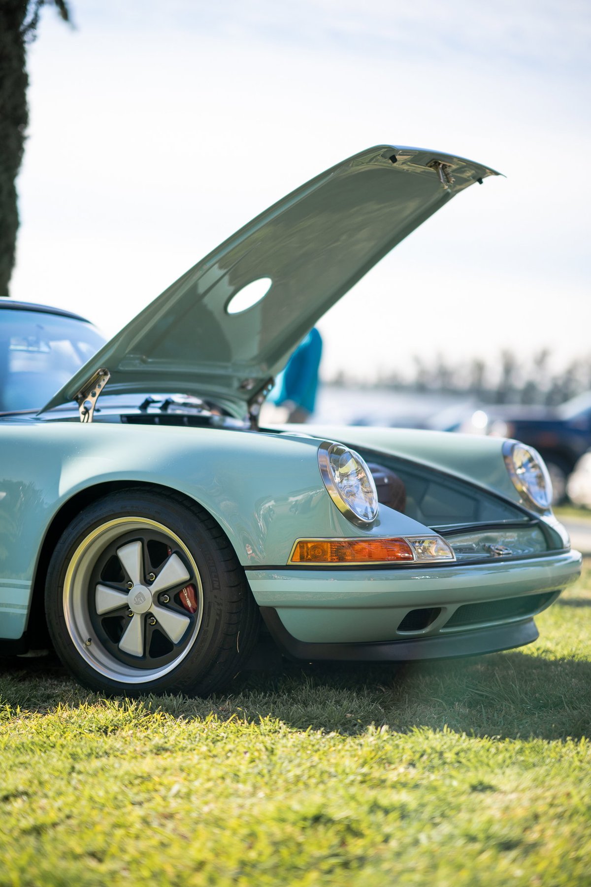 Light blue Porsche 911 with hood open, detailed classic rims, parked on grass.