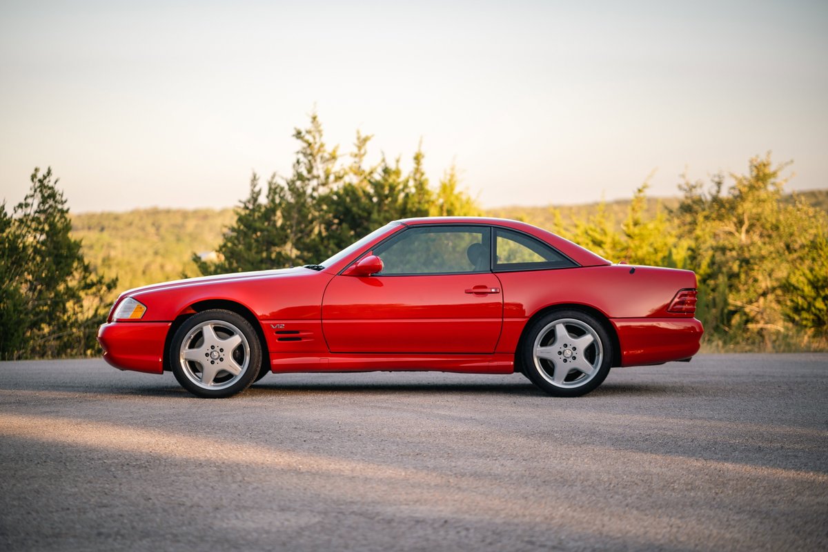 Red 2001 Mercedes-Benz SL600 with sport rims parked on asphalt road with trees in the background.