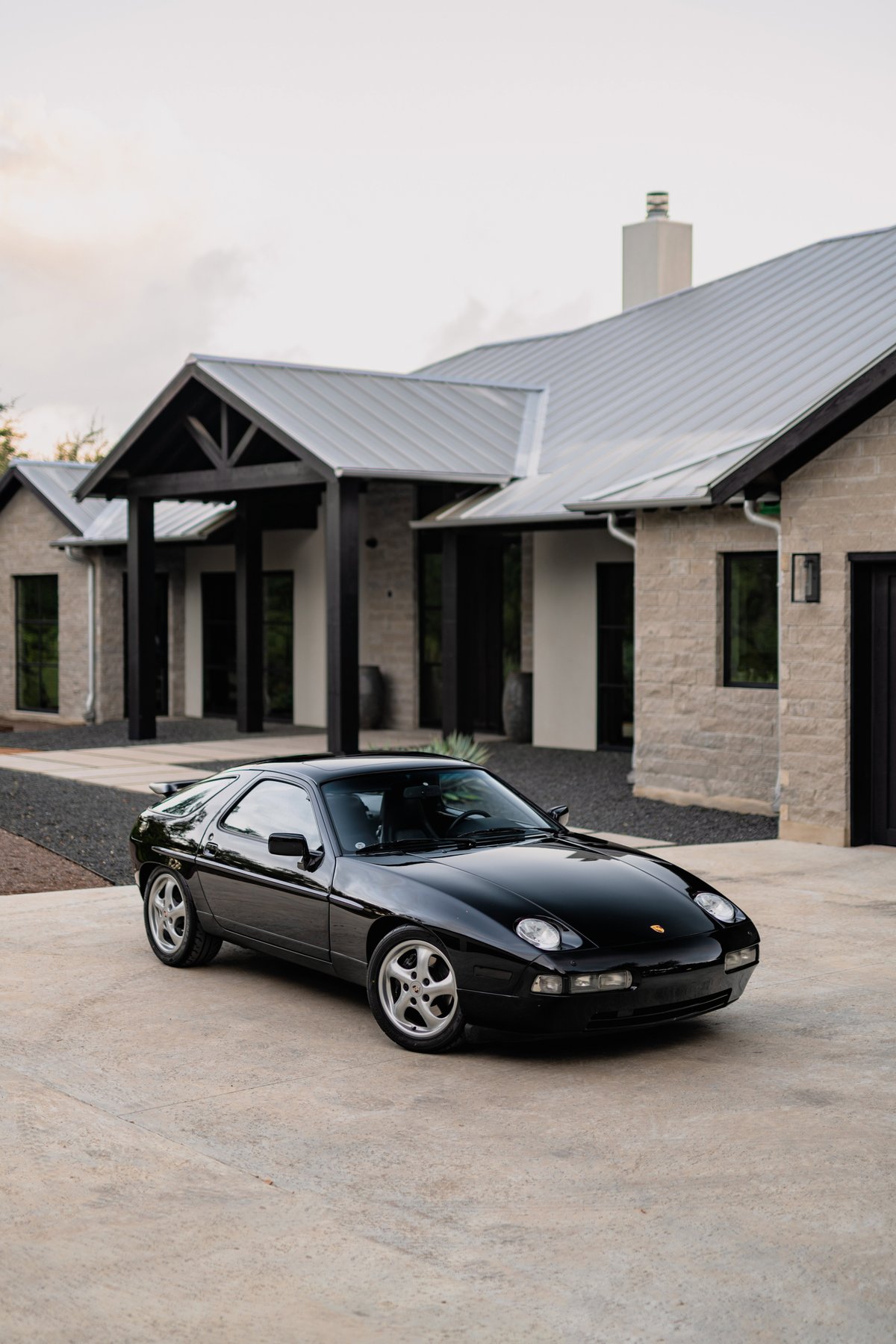 Black 1984 Porsche 928 S4 with silver five-spoke rims, parked in front of a modern stone house.