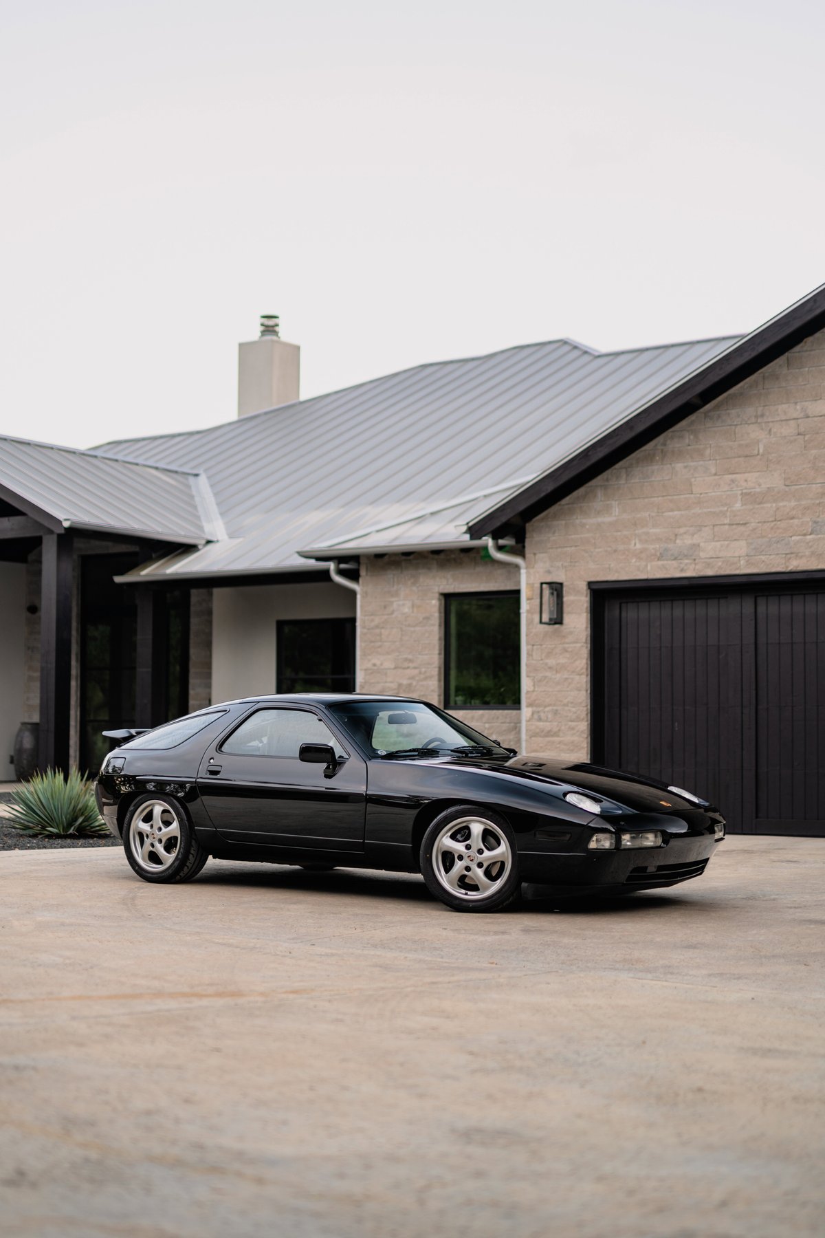 Black 1984 Porsche 928 S4 with silver cup style rims parked in front of a modern home.