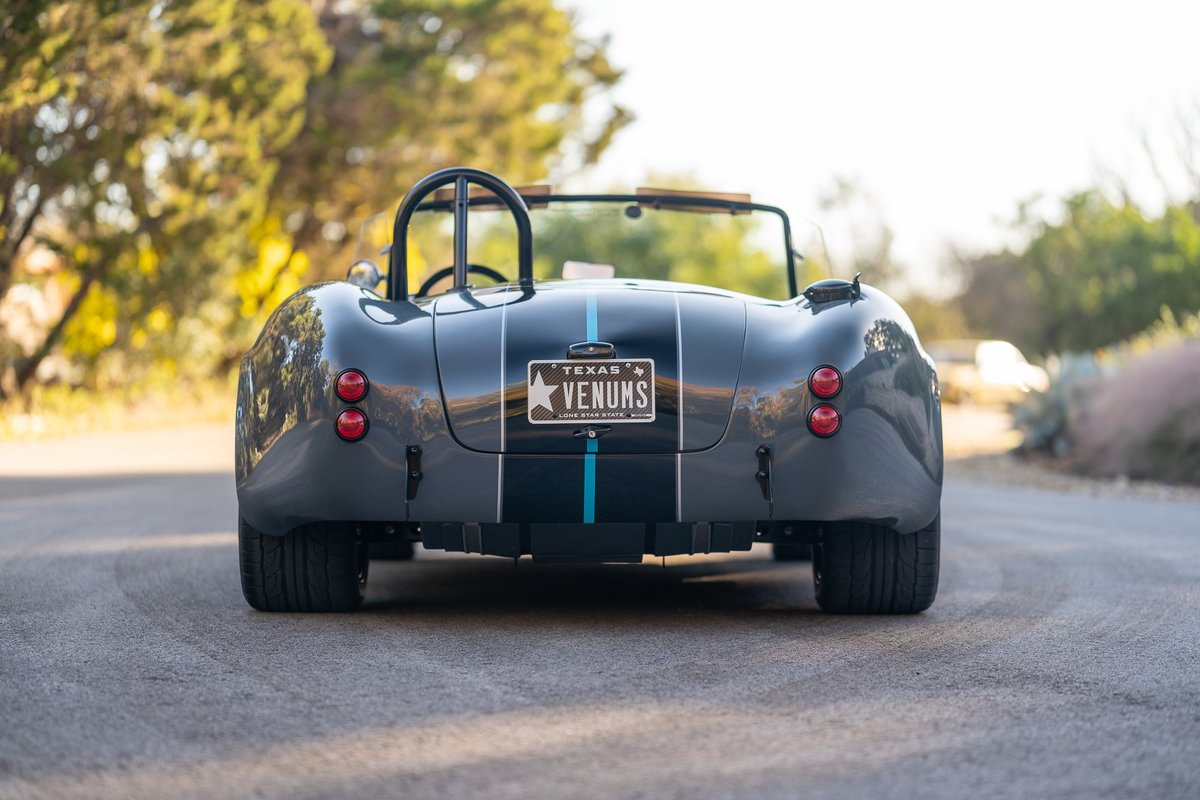 Rear view of a gray 1965 Backdraft Cobra with blue racing stripes and chrome roll bar.