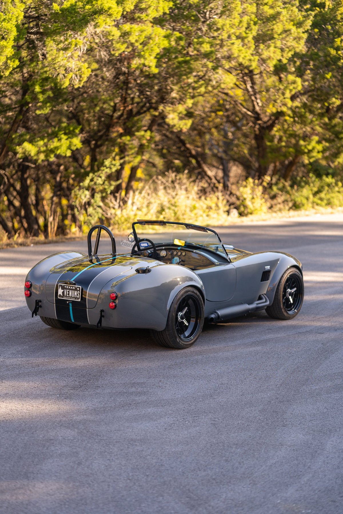 A gray Backdraft Cobra 427 with racing stripes and black rims sits on a paved road.