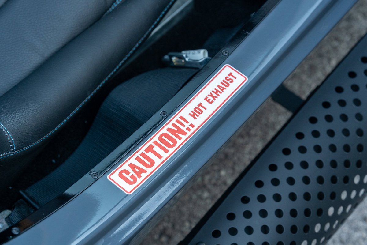 Close-up on a red and white "CAUTION!! HOT EXHAUST" decal on the gray door trim of a 1965 Backdraft Cobra.