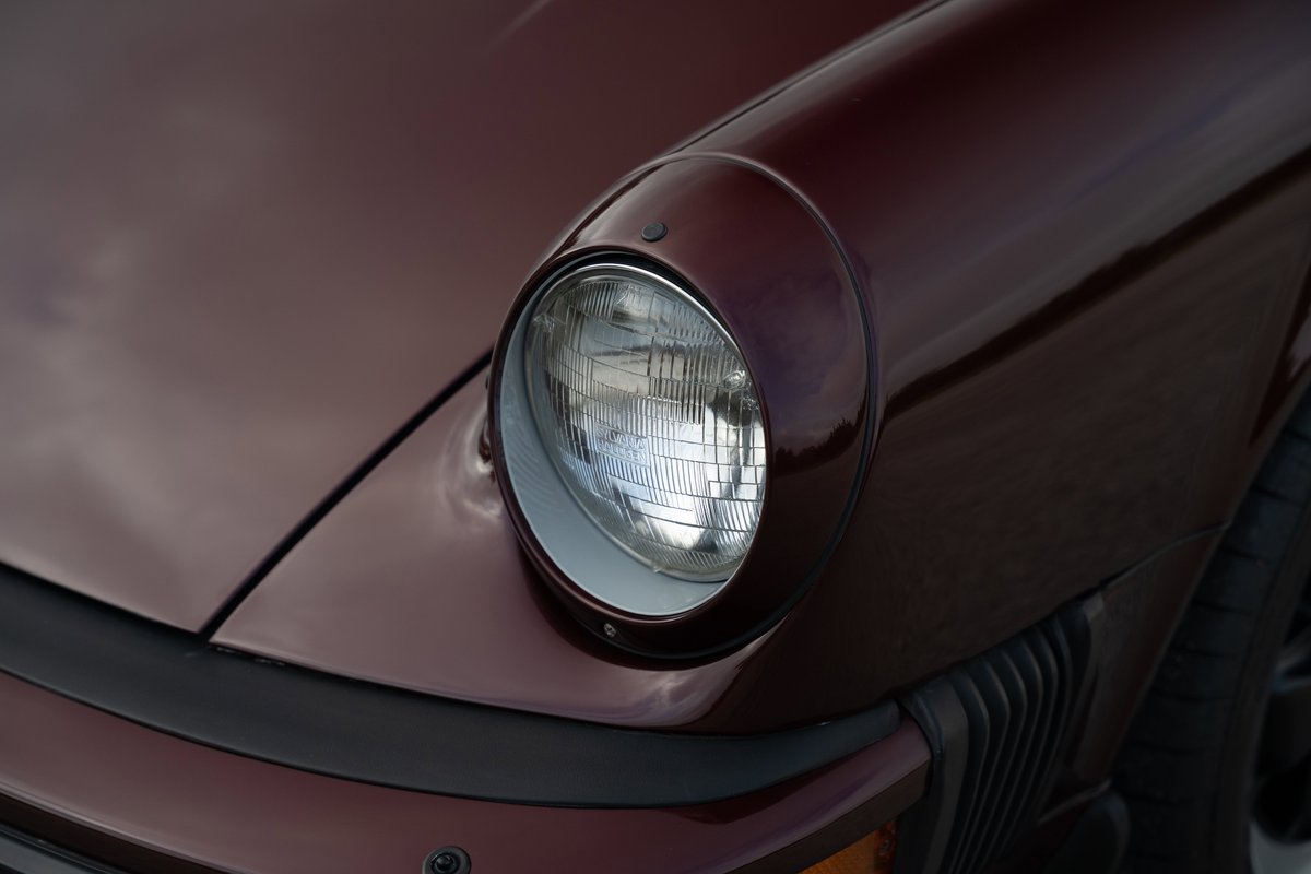 Ruby red metallic 1983 Porsche 911 SC Cabriolet headlight detail with chrome trim and black bumper.