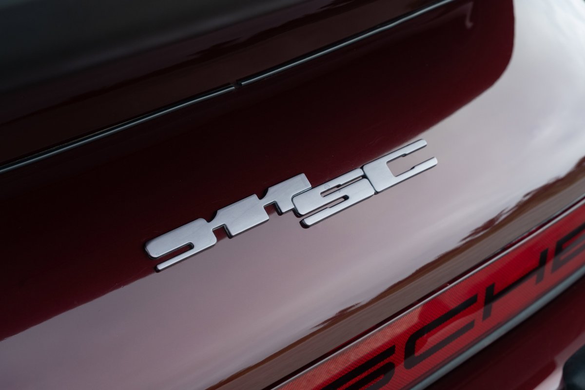Close-up of a silver "SC" badge on the rear decklid of a ruby red 1983 Porsche 911SC Cabriolet.