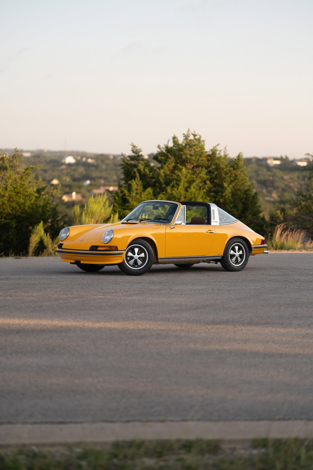A yellow 1973 Porsche 911S Targa with classic Fuchs wheels parked on an asphalt road.