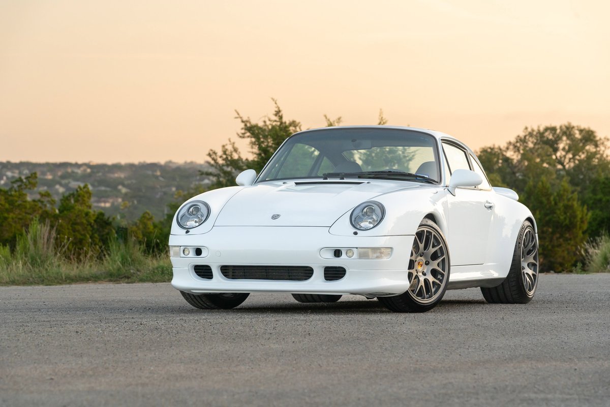 White 1997 Porsche 993 C4S with multi-spoke alloy rims parked on pavement with trees and hills.