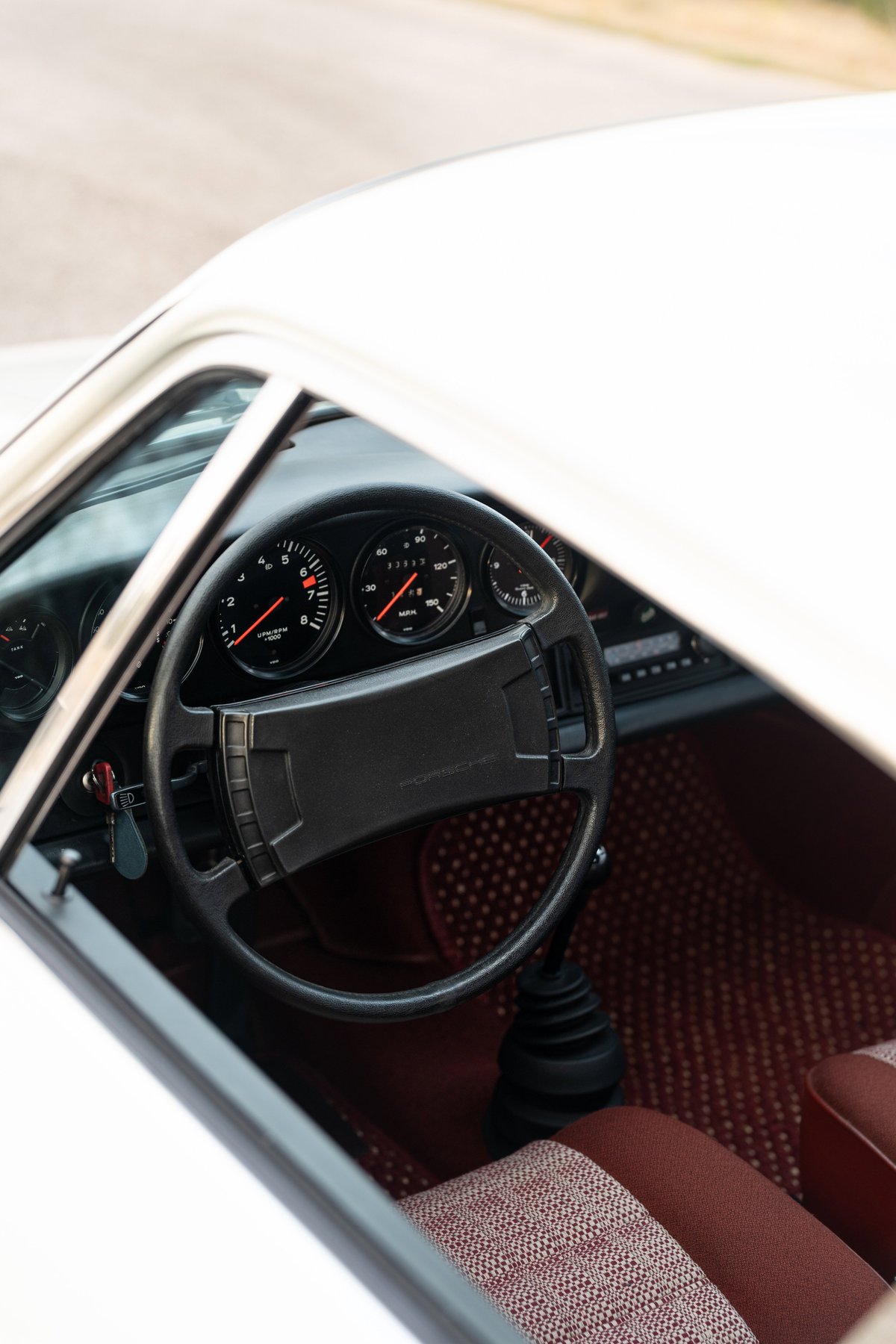 Interior of a white 1974 Porsche 911 Coupe, featuring a black steering wheel, red tartan seats, and a five-speed manual shifter.