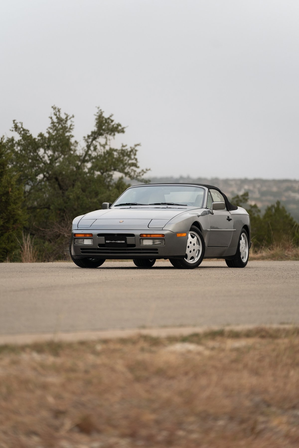 A grey 1990 Porsche 944 S2 Cabriolet with multi-spoke rims parked on a road.