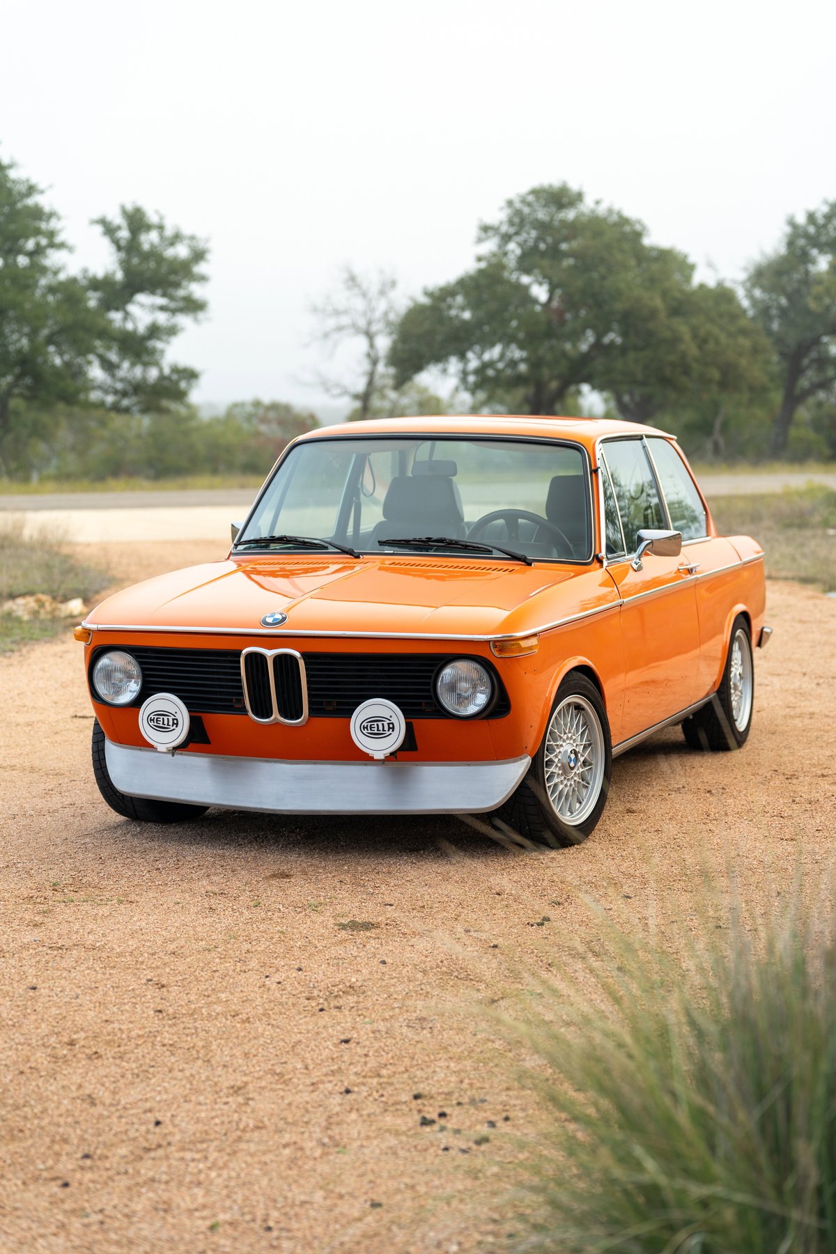 Orange 1976 BMW 2002 with chrome bumpers, Hella lights, and mesh rims.