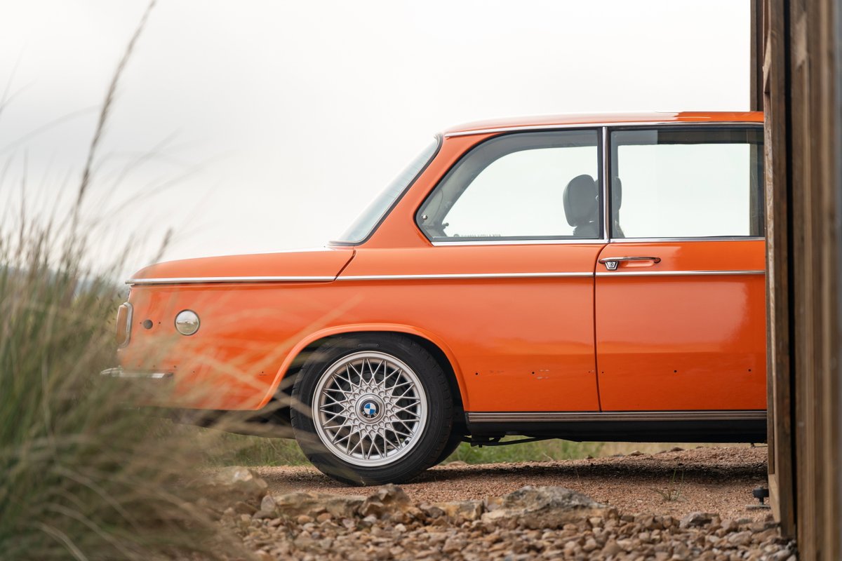 Side profile of an orange 1976 BMW 2002 with classic silver alloy rims.