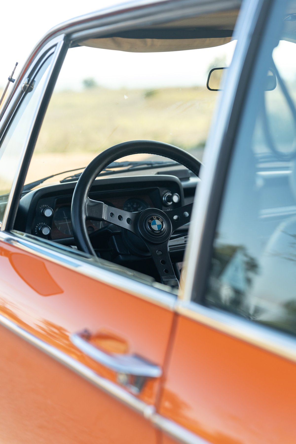 Interior of an orange 1976 BMW 2002. Features vintage steering wheel with BMW logo and chrome door handle.