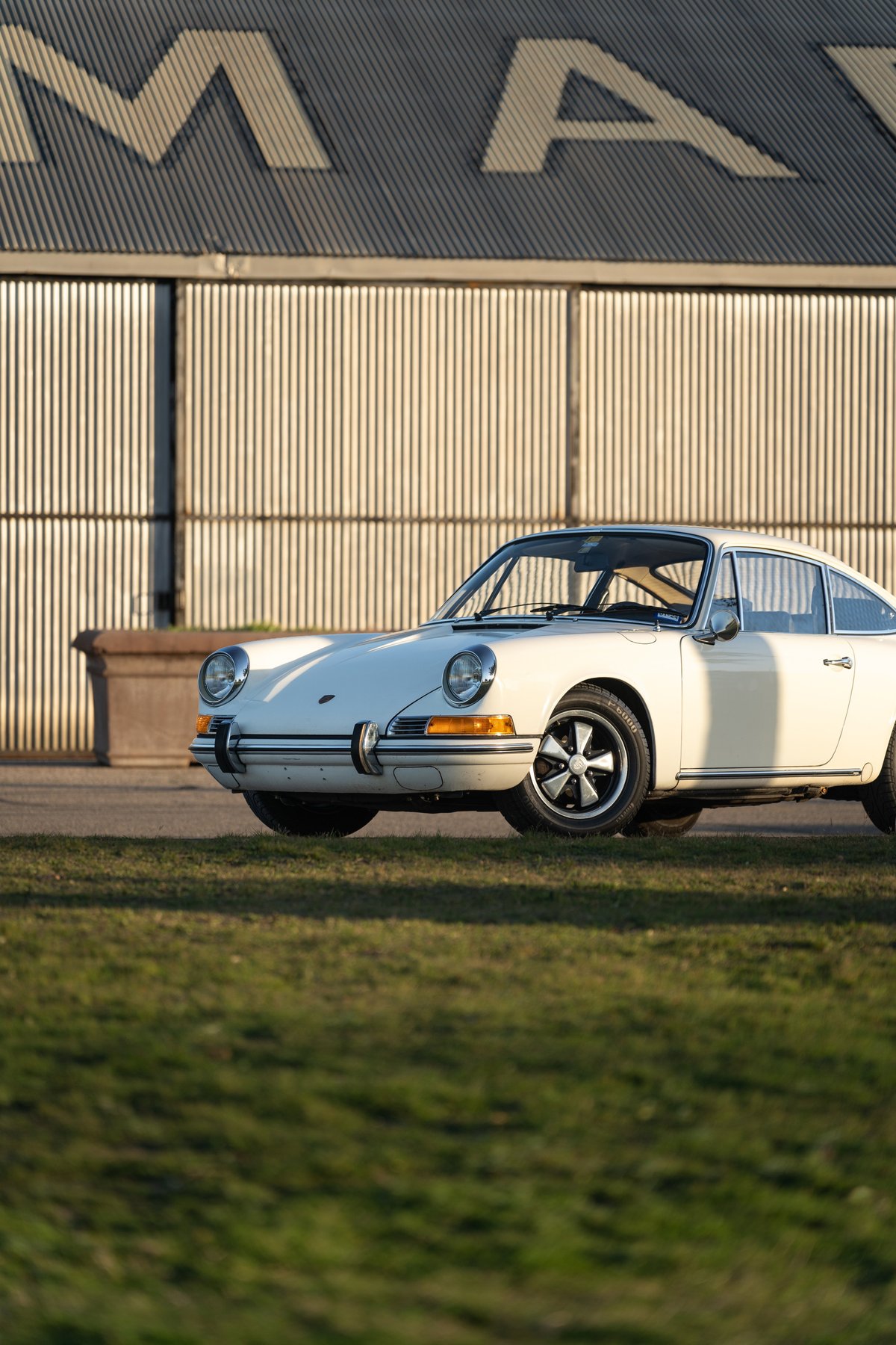 A white 1970 Porsche 911T Coupe with Fuchs rims parked on grass in front of a building.