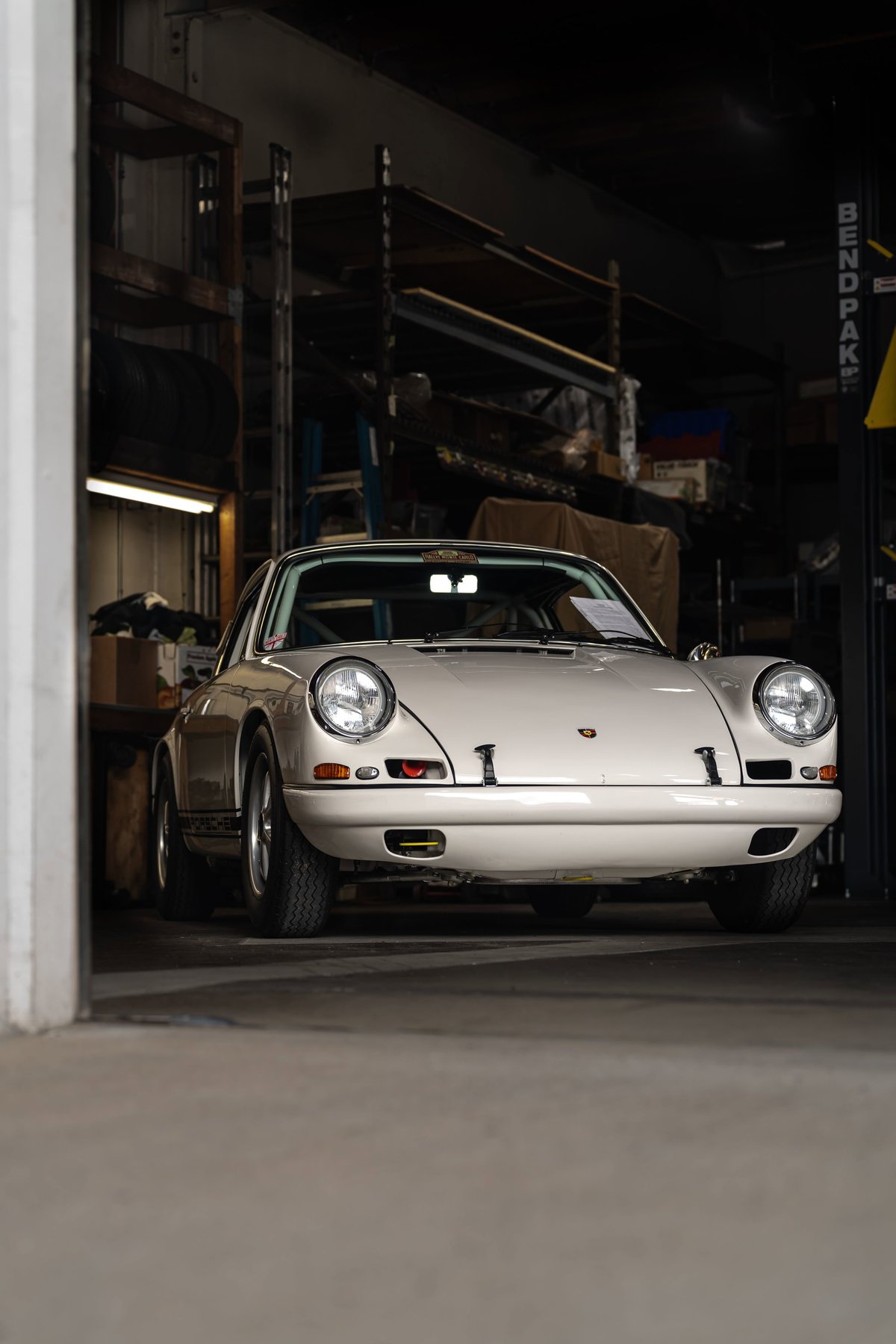 A white 1972 Porsche 911 Carrera RS 2.7 sits in a workshop, showcasing its chrome trim and classic Fuchs rims.