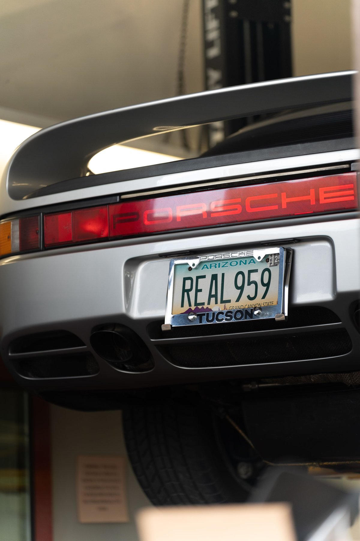 Silver Porsche 959 with "REAL 959" license plate, showcasing its rear spoiler and taillights.
