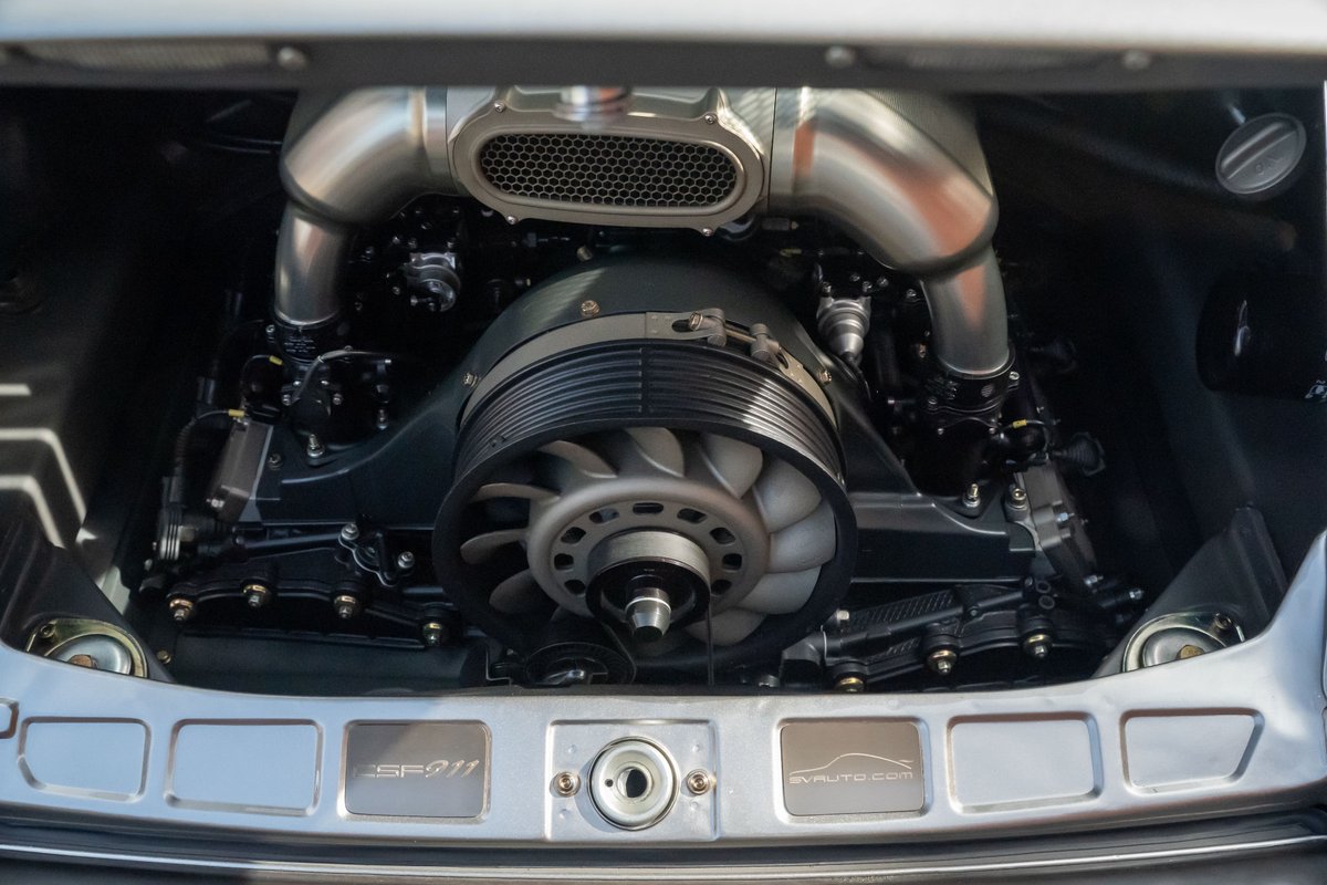 Close-up of a silver Porsche 911 engine with polished intake pipes and a visible fan.