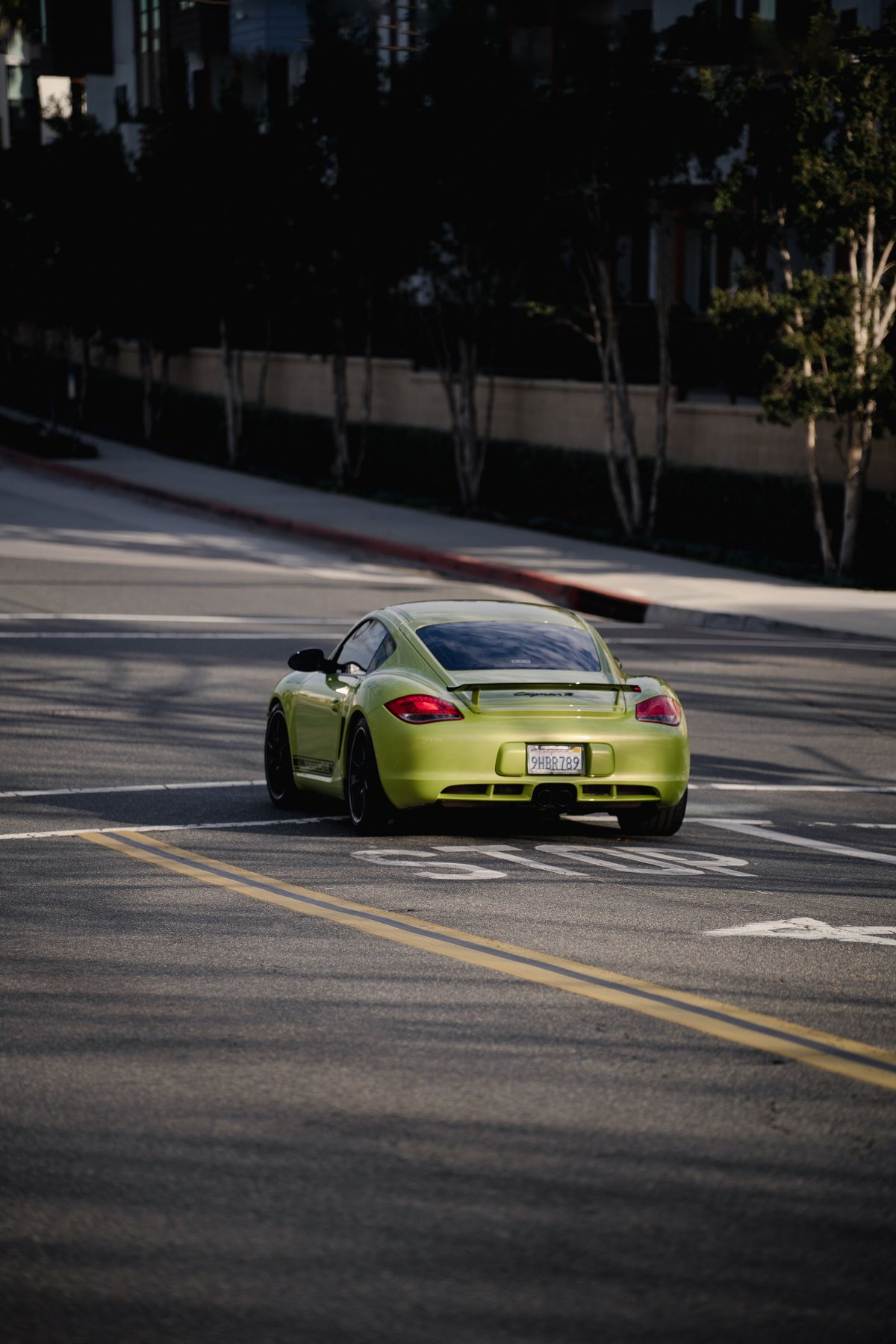 Rear view of a lime green Porsche Cayman S with black rims and a spoiler driving on the road.