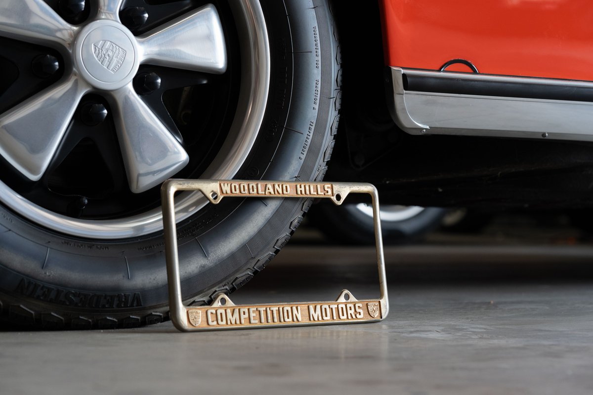 Close-up of a vintage Porsche's Fuchs rim and orange paint with a "Competition Motors" license plate frame.