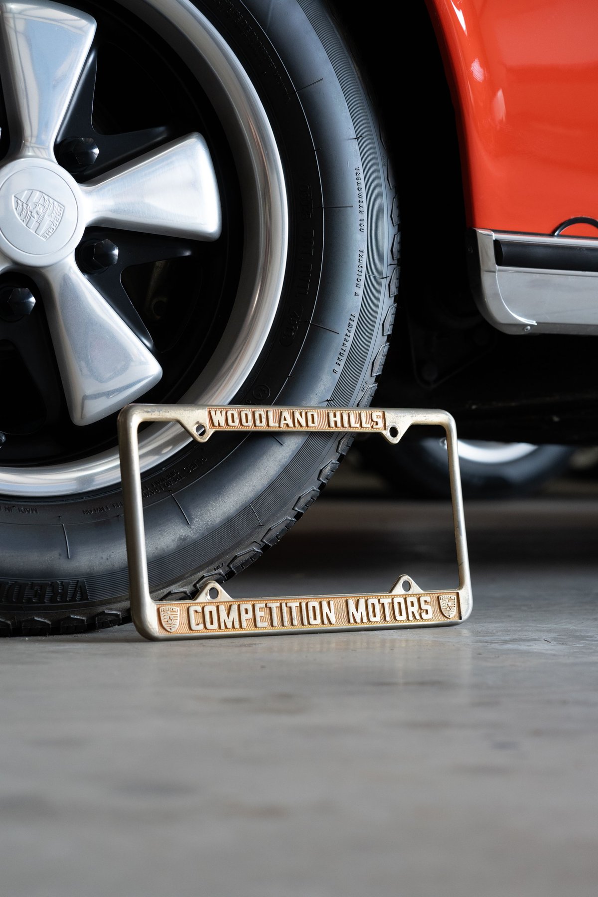 Porsche Fuchs rim and chrome trim on orange classic car with "Competition Motors" license plate frame.