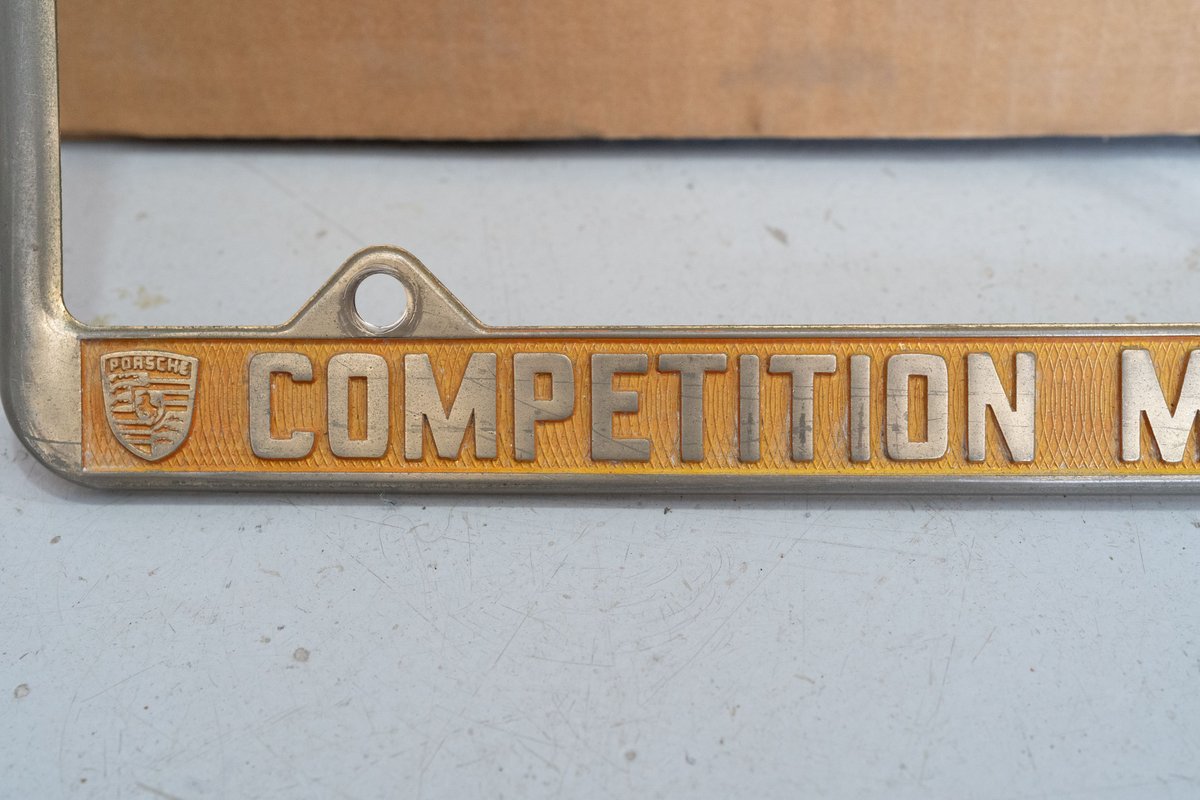 Close-up of a vintage Porsche Competition Motors license plate frame with gold trim and emblem.
