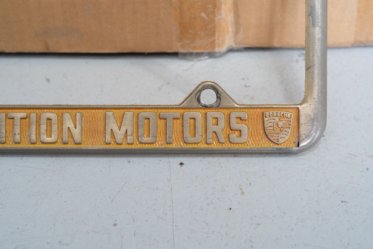 Close-up of a silver license plate frame with gold trim and "Competition Motors" text, featuring a Porsche crest emblem.