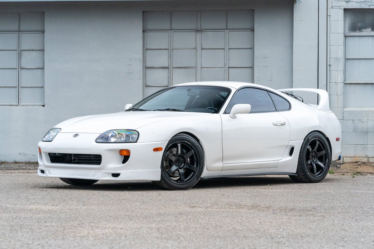 White 1994 Toyota Supra with aftermarket front bumper, rear spoiler, and black rims.