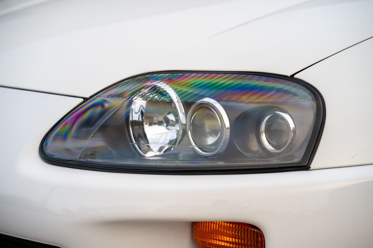 Closeup of a white 1994 Toyota Supra headlight with rainbow prism effect on the lens.