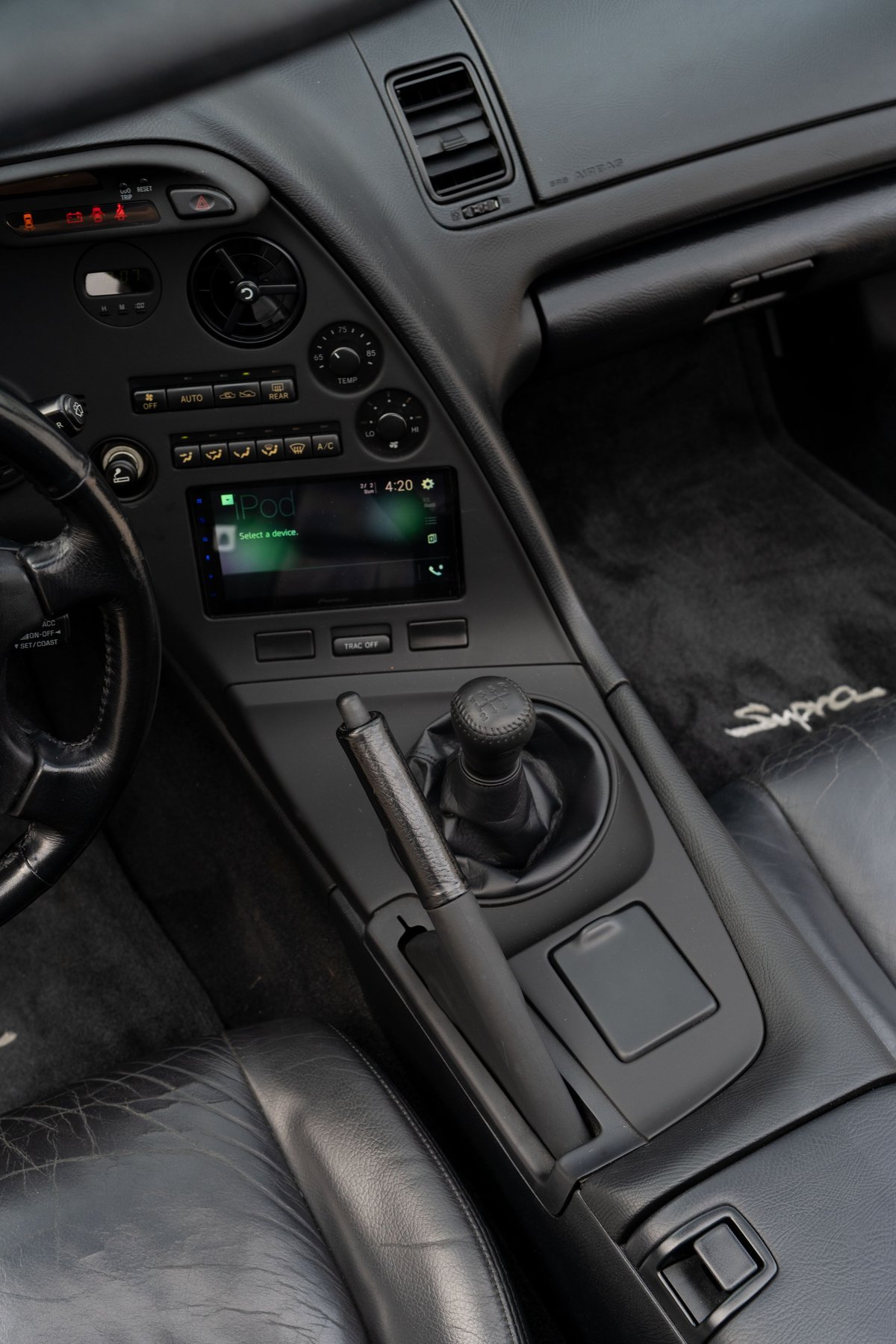 Black leather interior of a 1994 Toyota Supra, featuring a 5-speed manual shifter, modern infotainment screen, and "Supra" floor mat.