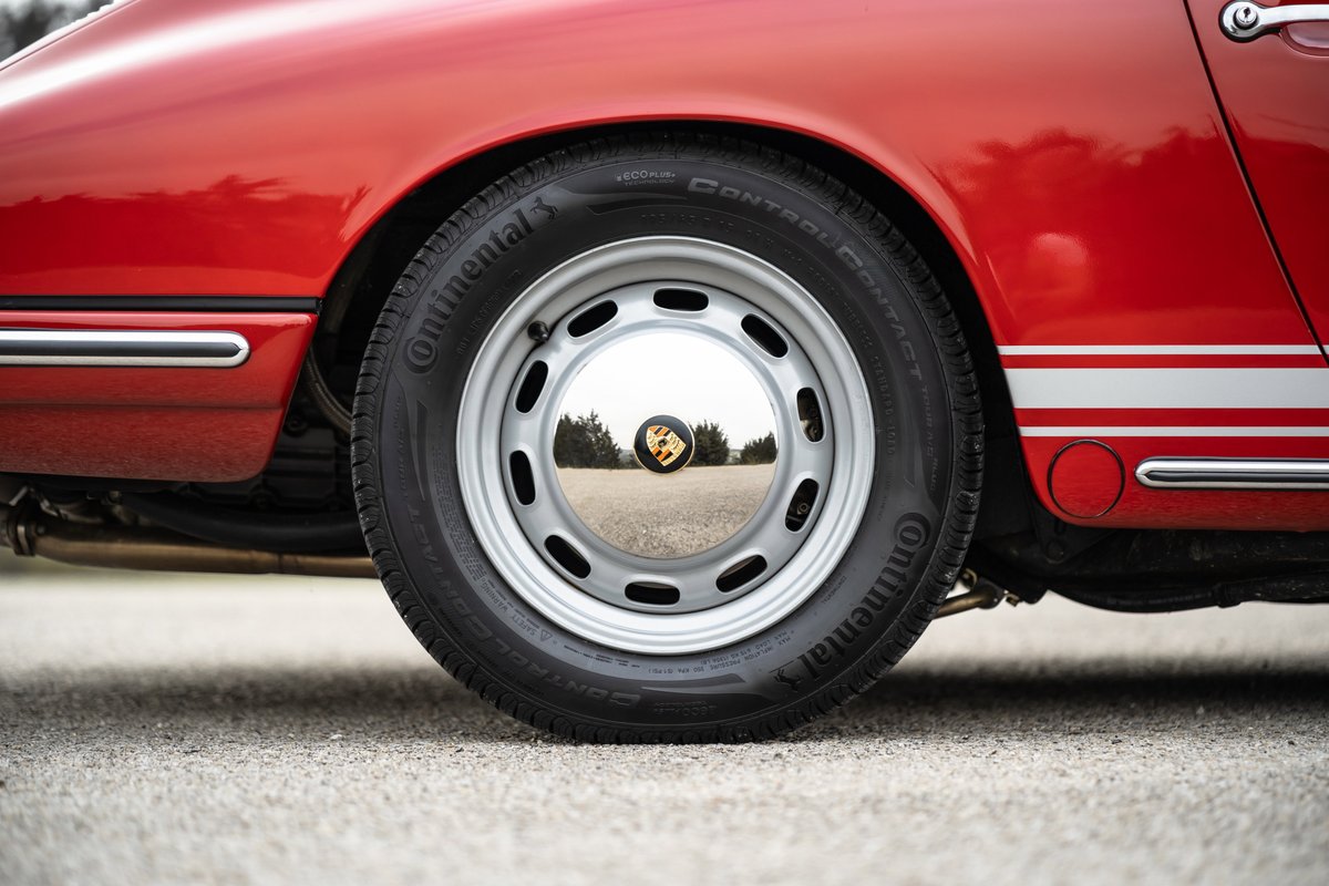 Rear wheel of a red 1967 Porsche 912 coupe with chrome trim and a Porsche crest hubcap.