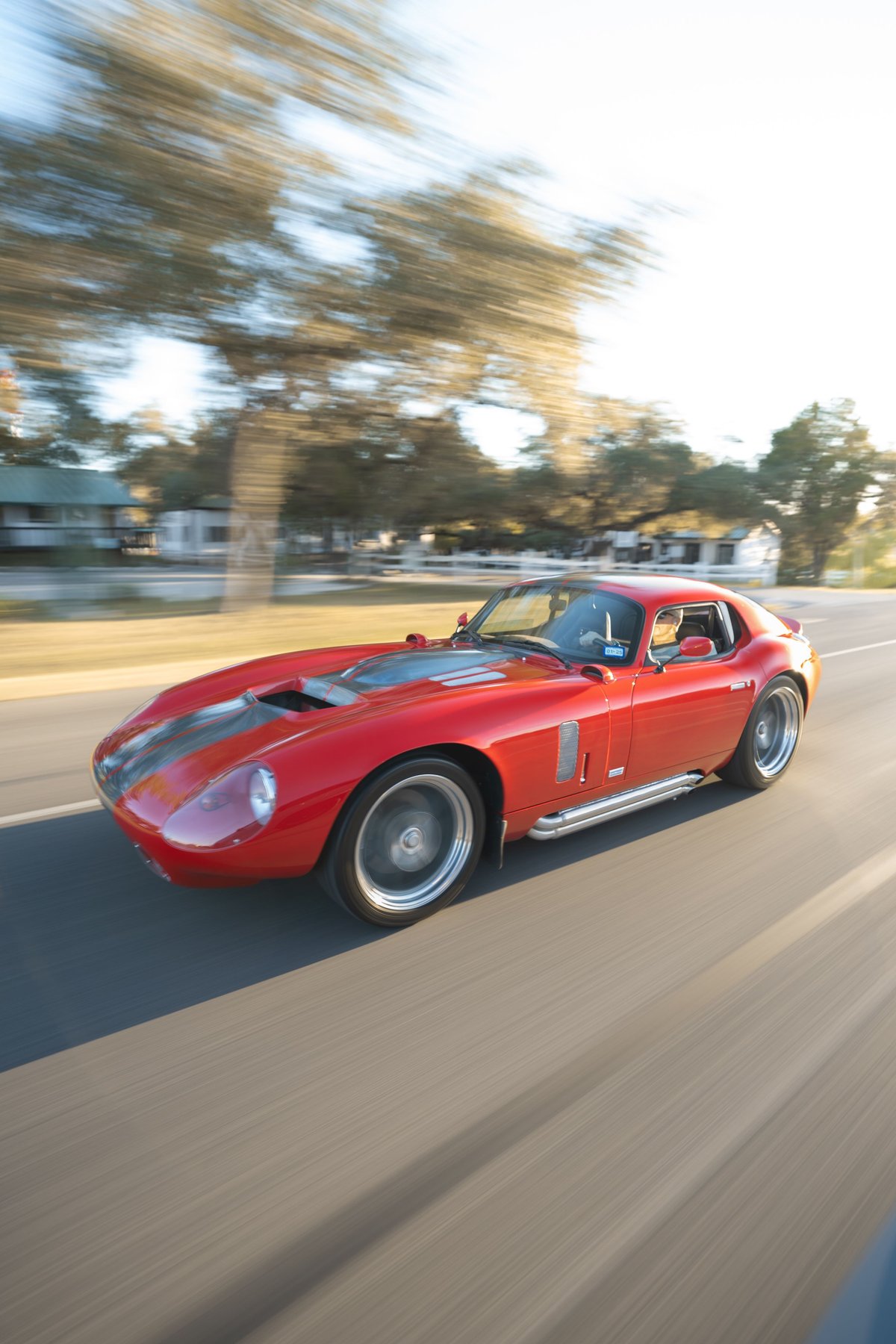 Red 1965 Superformance Shelby Daytona Coupe with dual gray racing stripes and chrome side pipes on alloy rims.